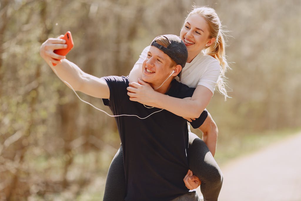 A joyful couple taking a selfie while enjoying a piggyback ride in a sunny park.