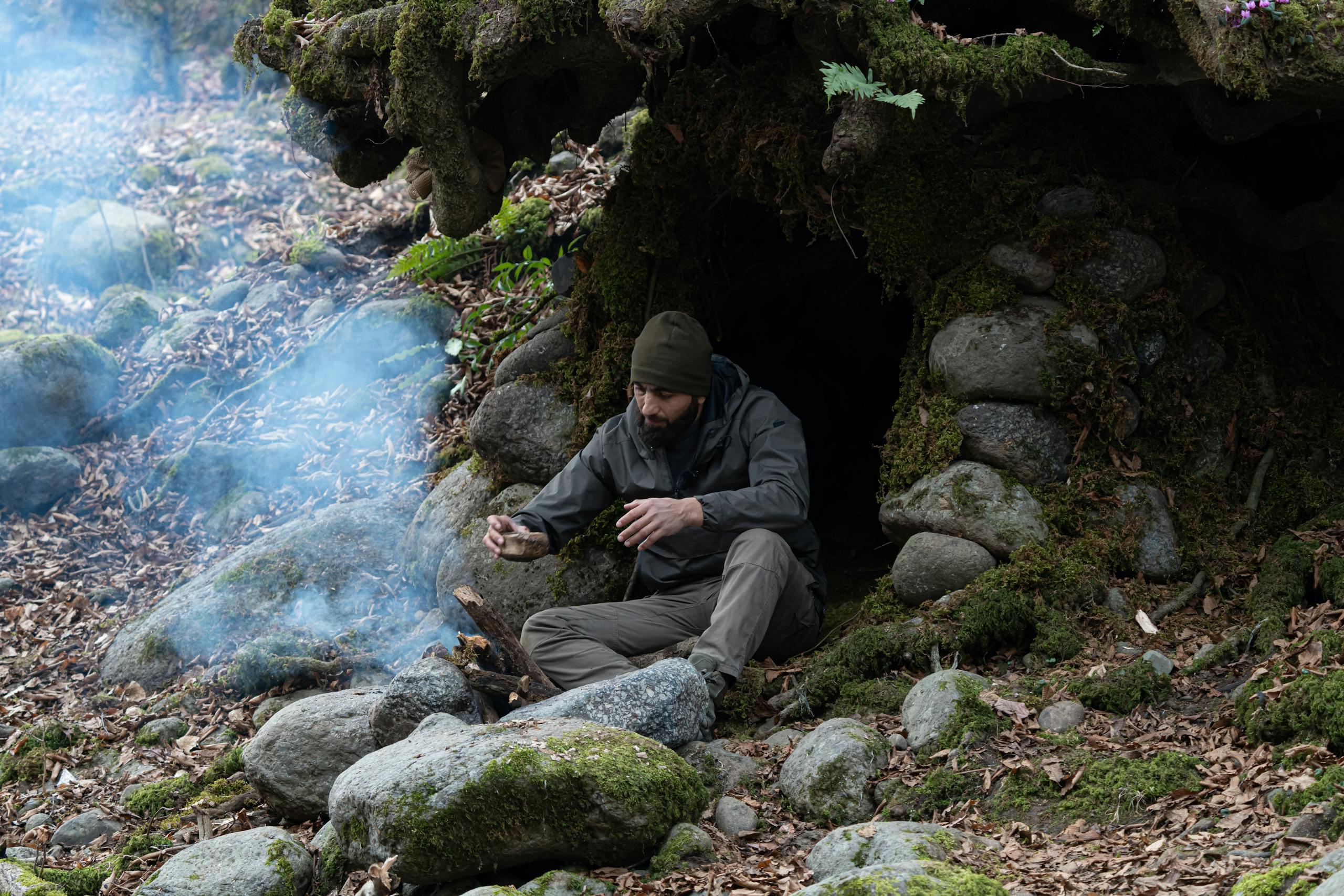 A man sits by a stone shelter making a fire in a forest setting with smoke.