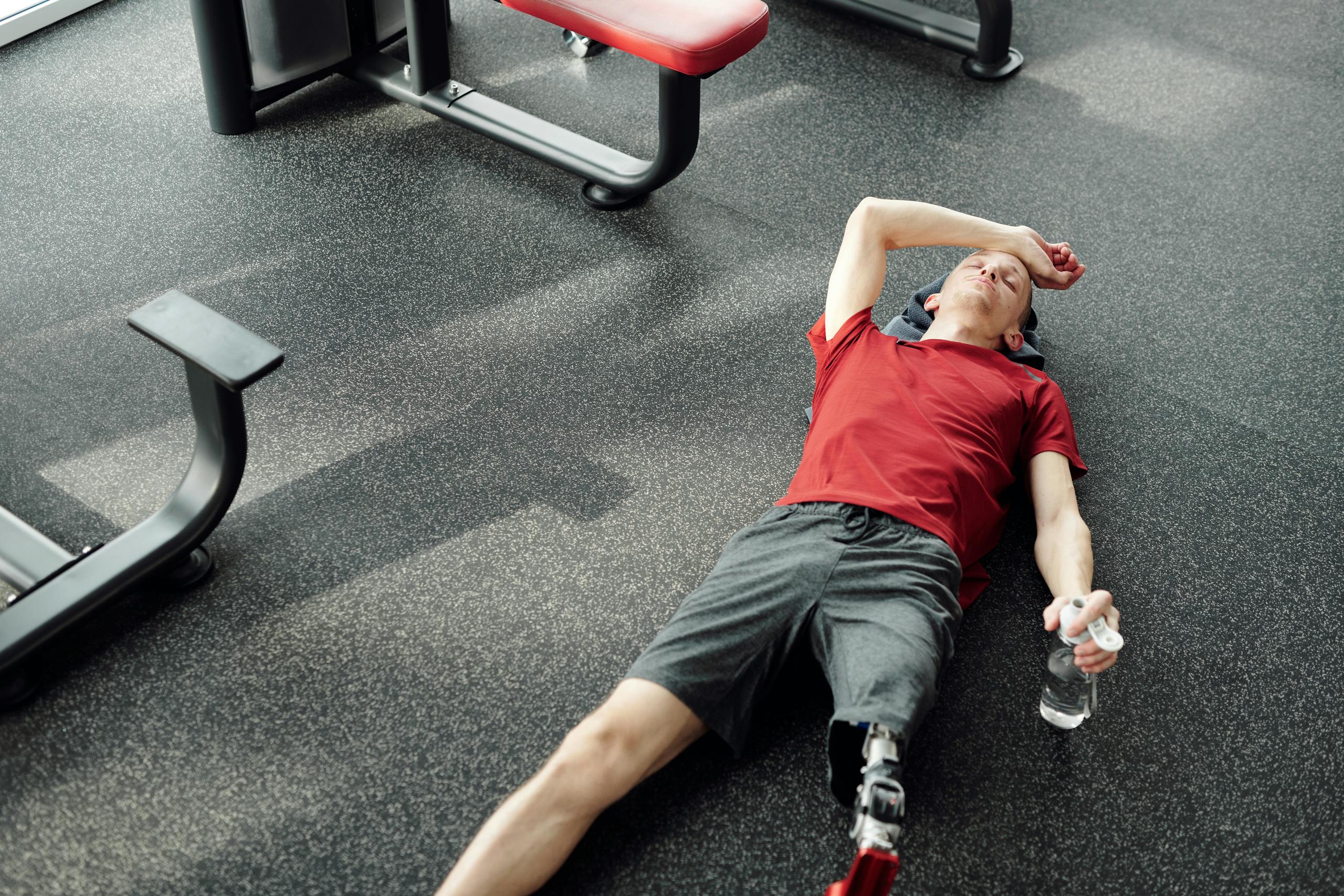 A man with a prosthetic leg rests on the gym floor, holding a water bottle after an intense workout.