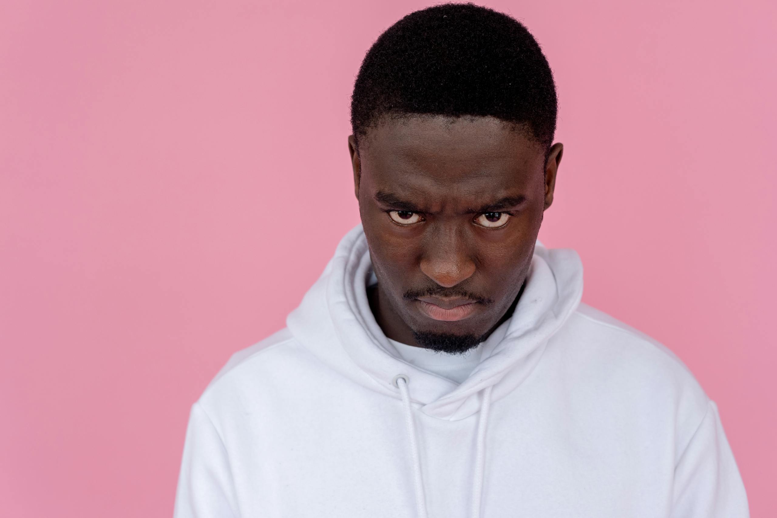 A man with a serious expression, wearing a white hoodie against a pink background.