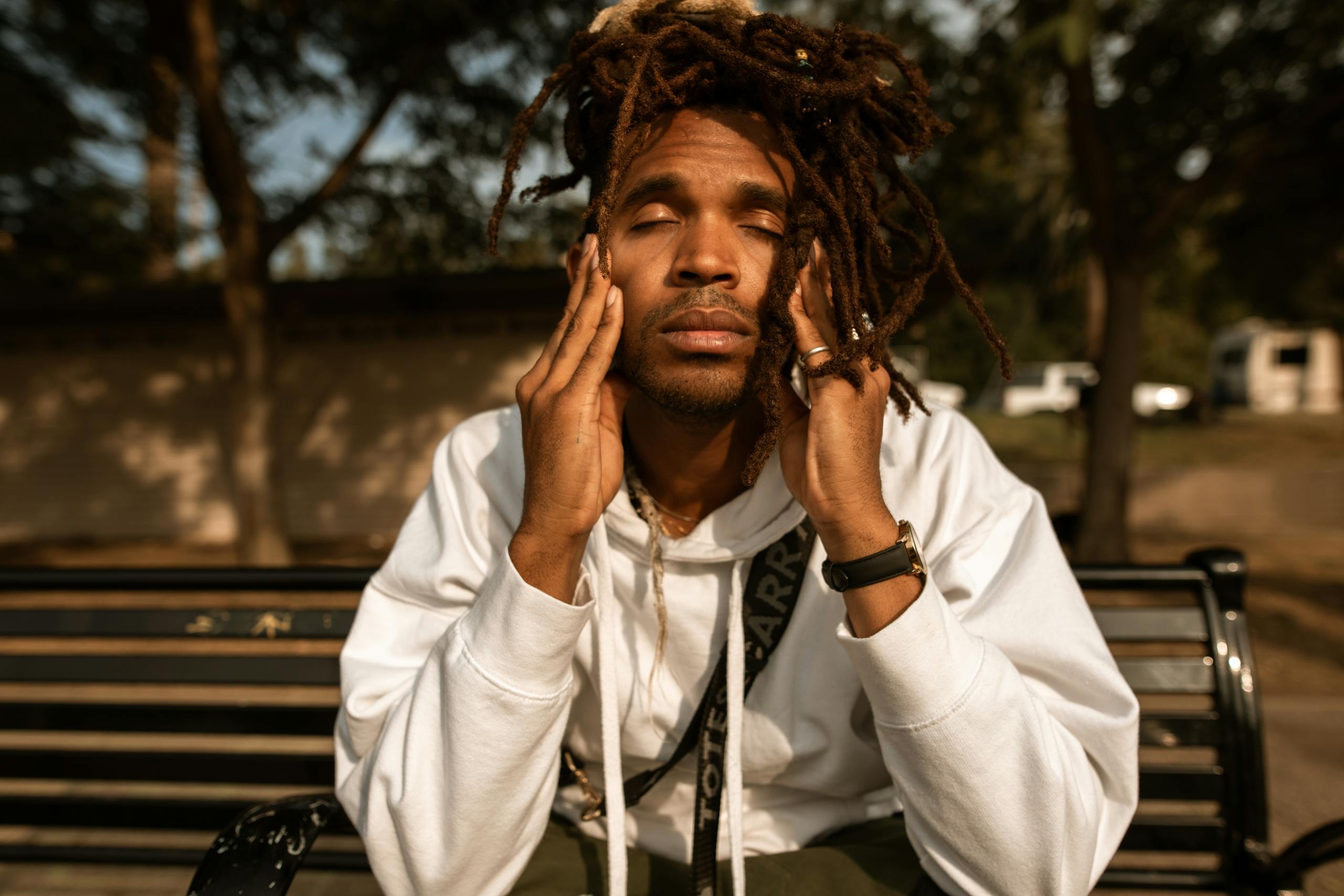 A man with dreadlocks sits on a park bench, contemplating with eyes closed.