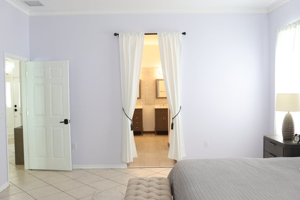 A minimalist bedroom interior featuring white curtains, soft lighting, and an open bathroom view.