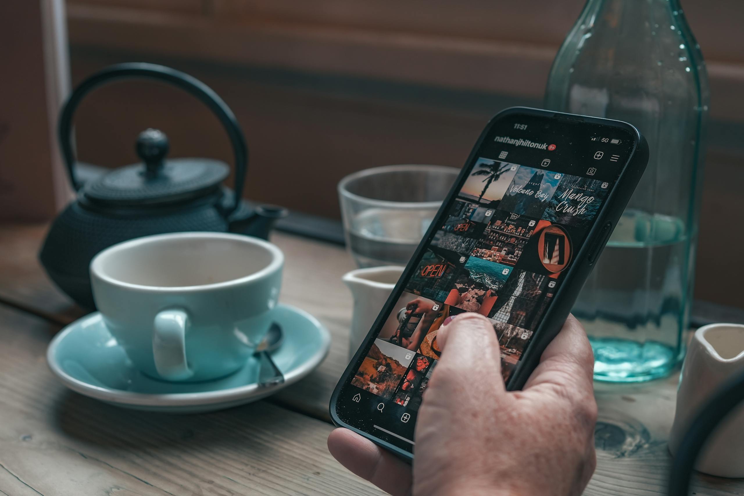 A person holds a smartphone browsing Instagram next to a teacup and teapot on a wooden table.