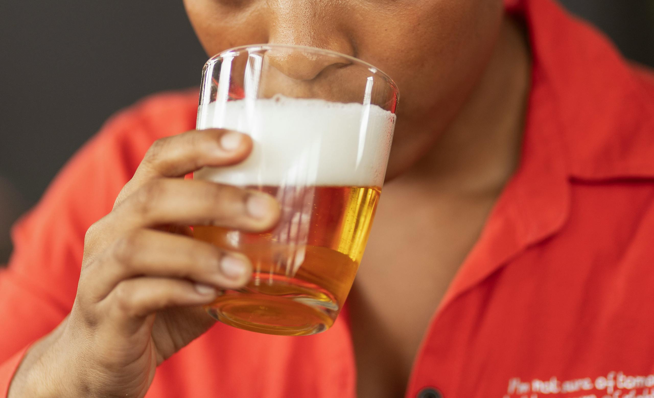 A person in a red shirt taking a sip of beer from a glass.