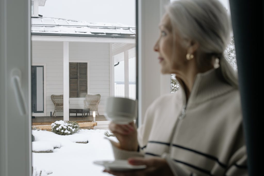 A senior woman holding a ceramic cup looks out a window at a snowy winter porch.