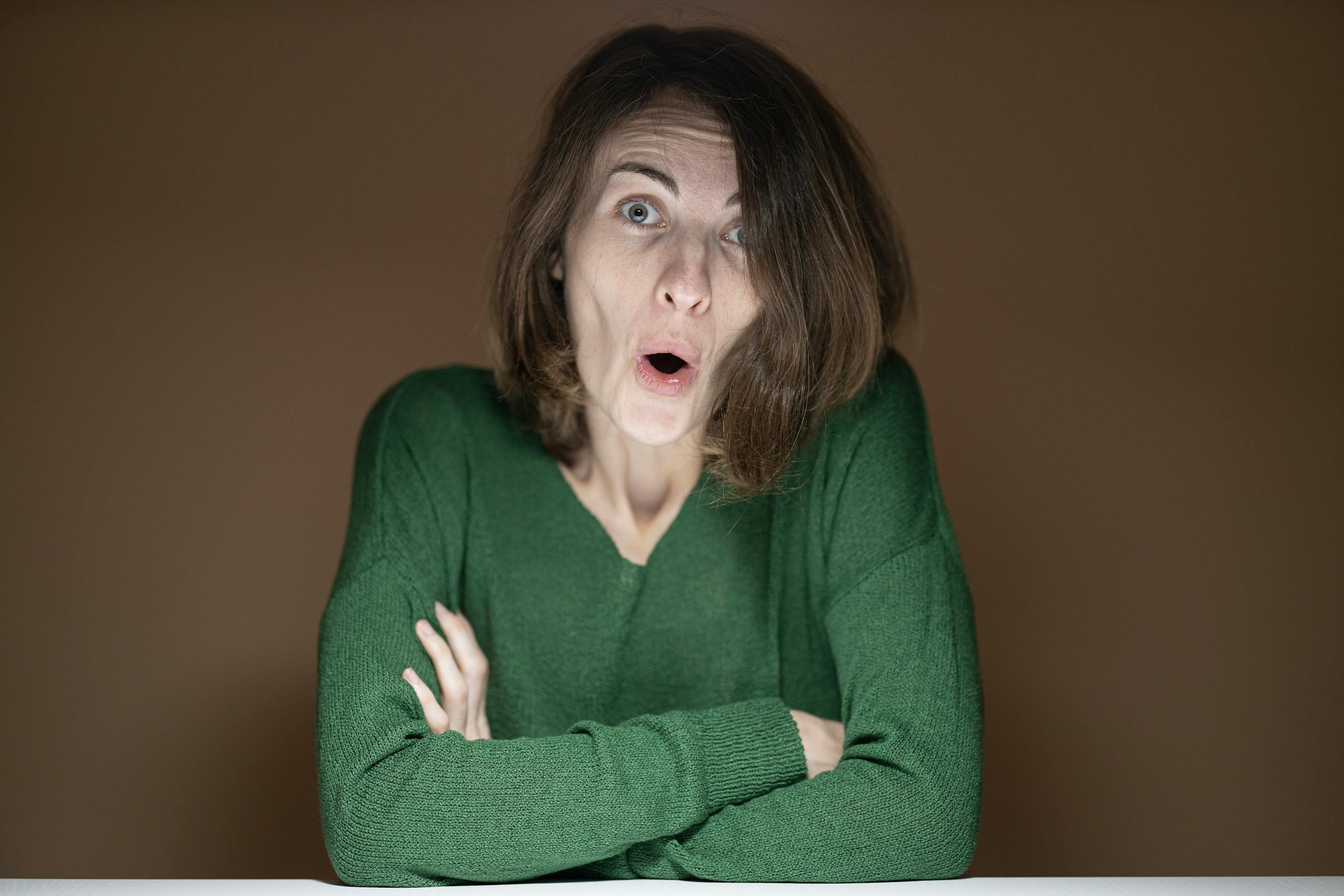 A shocked woman in a green sweater with a surprised expression poses against a neutral background.