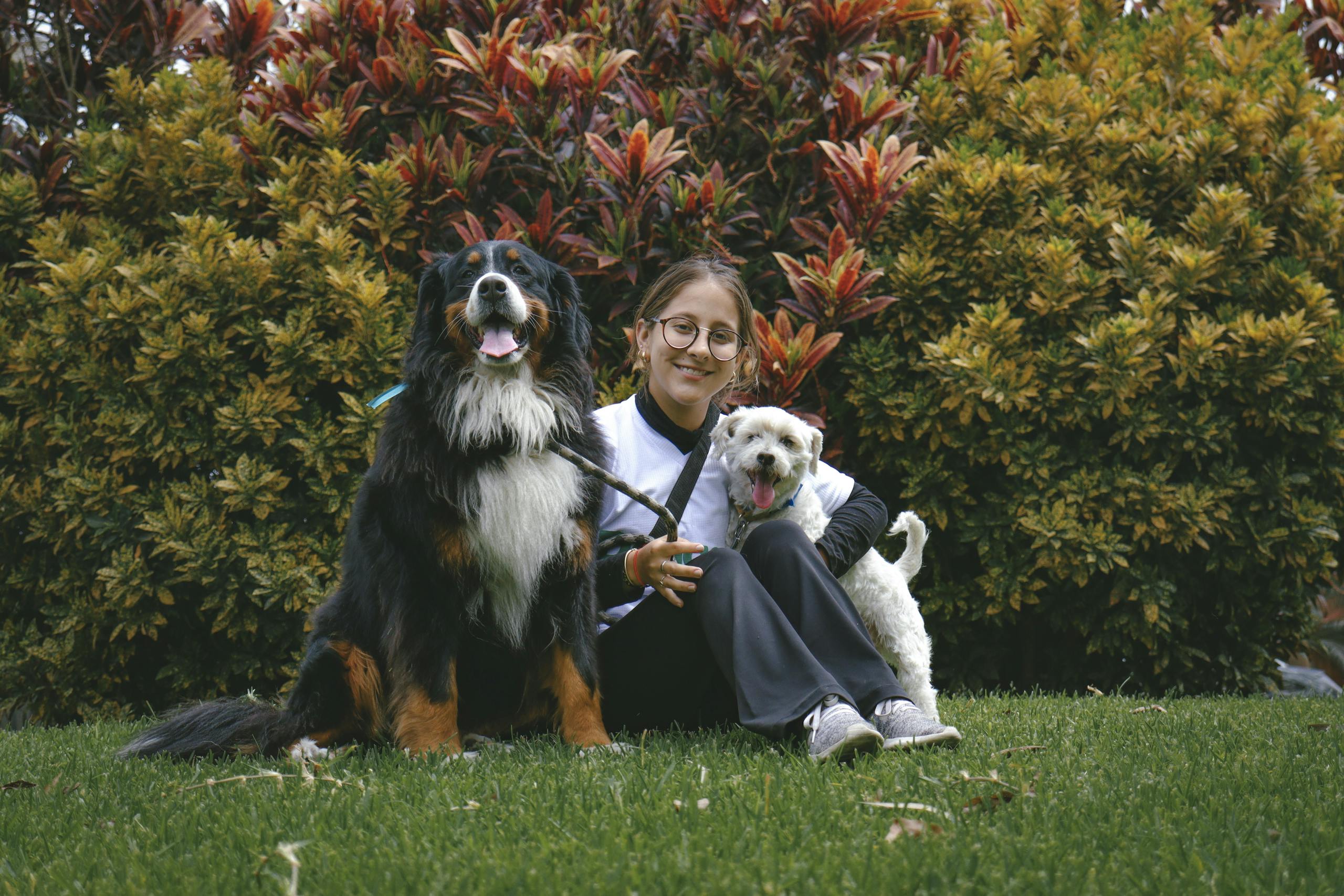 A smiling woman sits with two happy dogs in a lush garden, surrounded by colorful foliage.