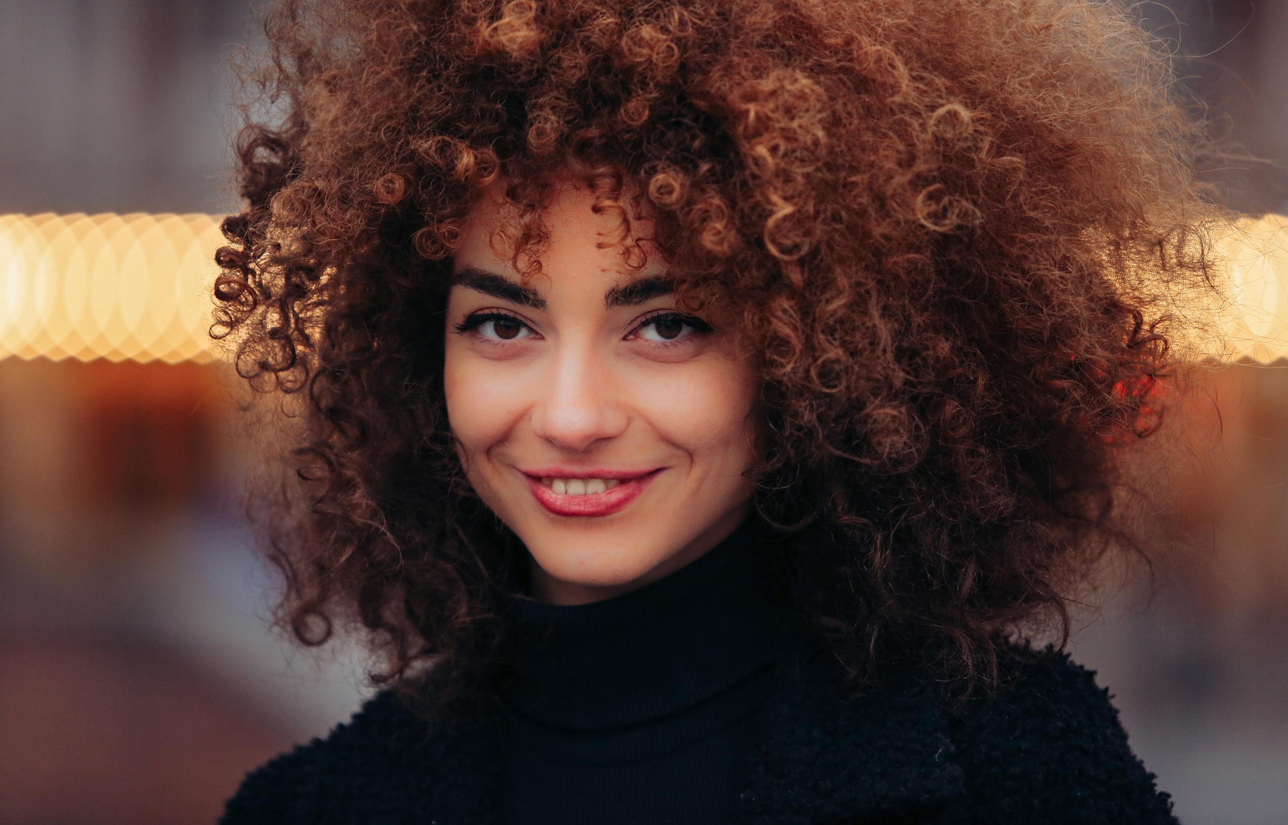 A stunning portrait of a woman smiling warmly, featuring natural curly hair and a stylish black outfit.