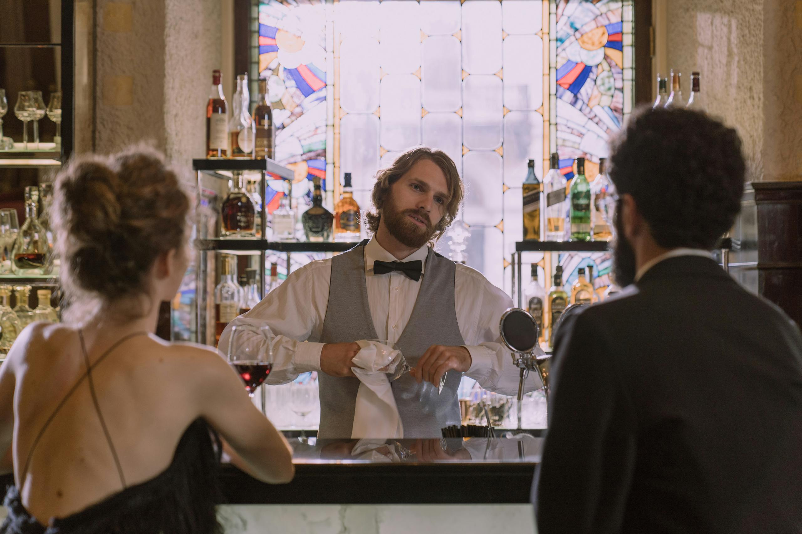 A stylish bartender serves drinks to a couple in a luxurious bar setting with stained glass details.