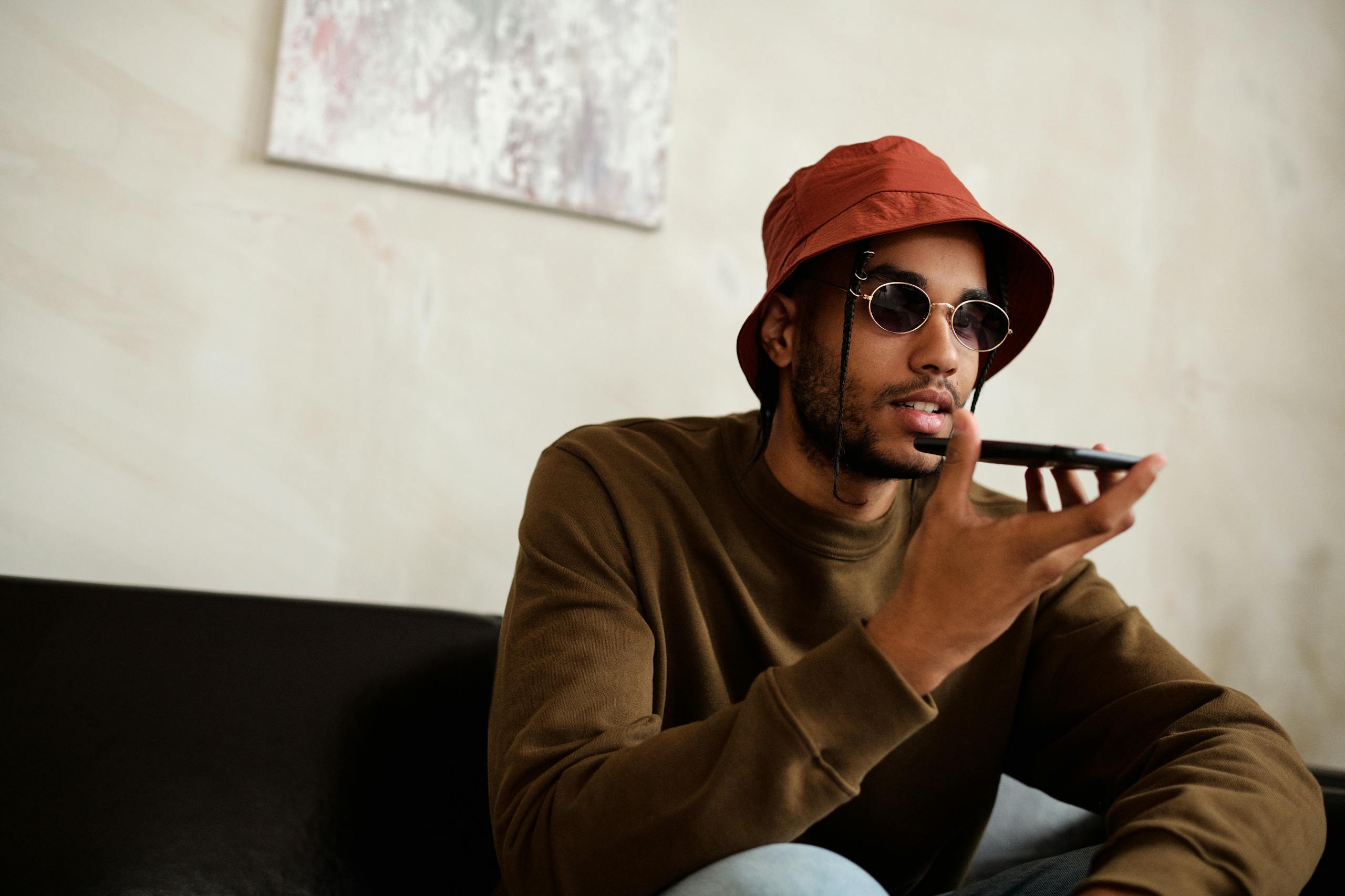 A stylish young man with sunglasses and a bucket hat using a smartphone for a voice call indoors.