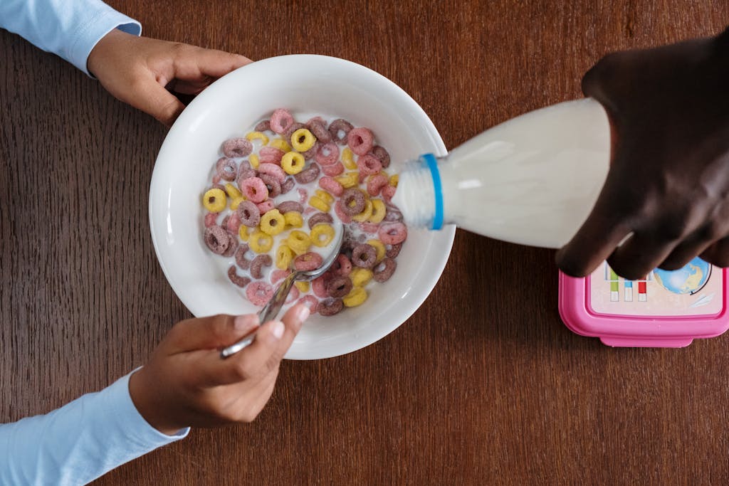 A top view of hands pouring milk into a bowl of colorful cereal on a wooden table.