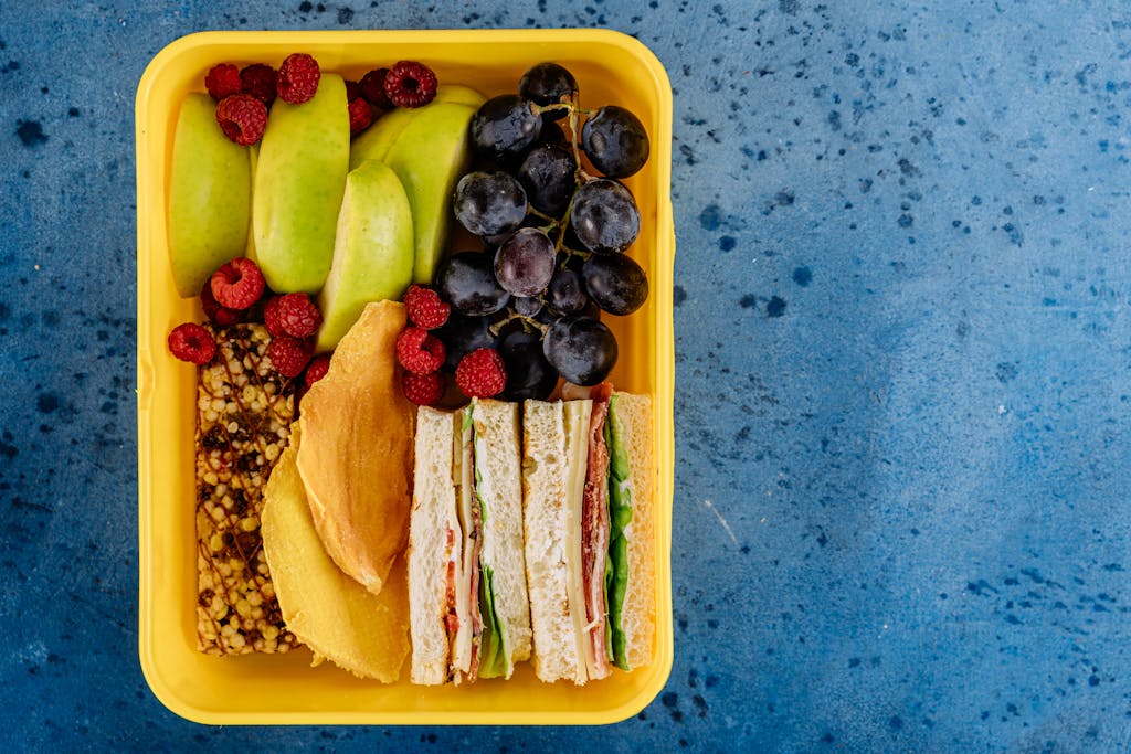 A vibrant lunchbox filled with sandwiches, apples, grapes, and raspberries on a blue background.