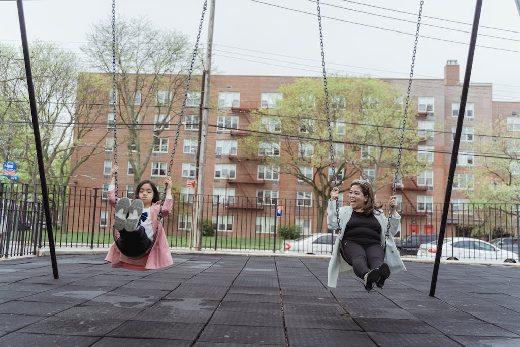 A woman and a girl swinging together in a city playground, enjoying a bright day outdoors.