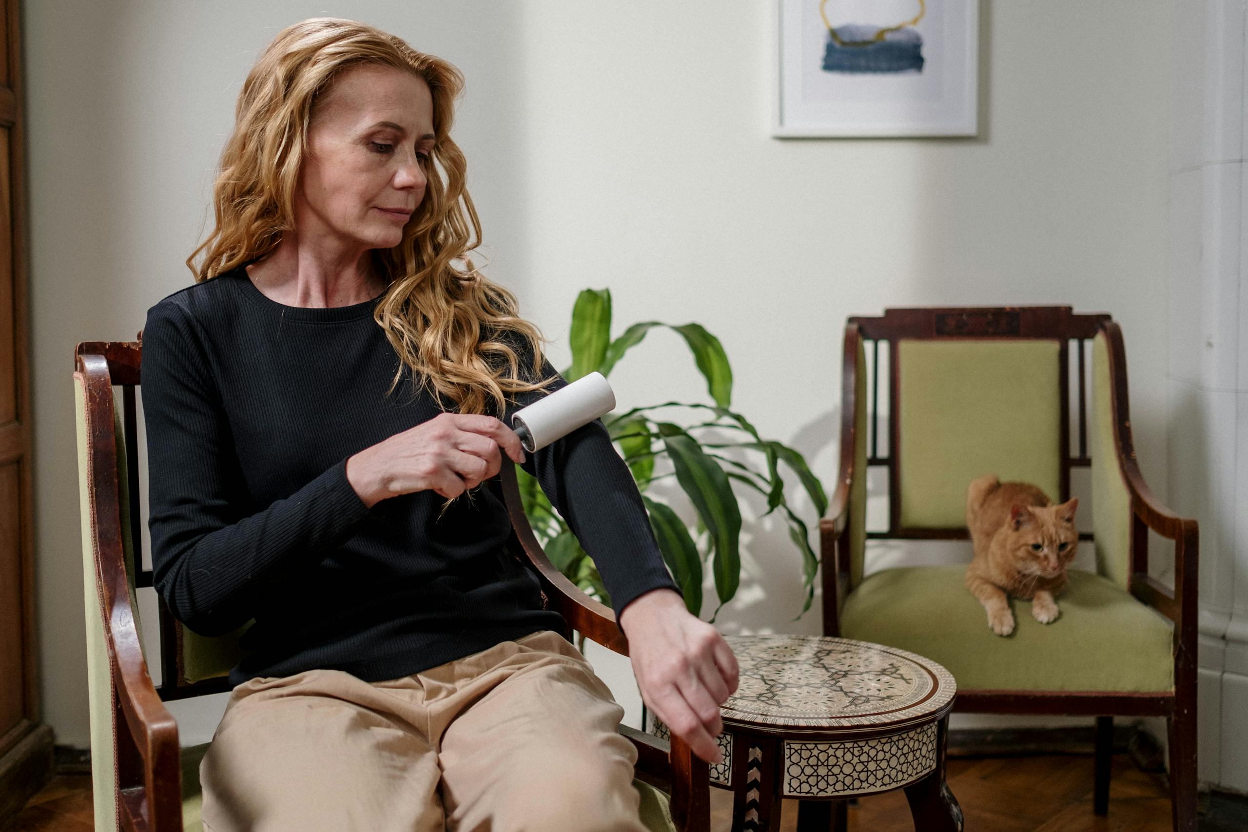 A woman cleaning her clothes with a lint roller while a ginger cat sits nearby, indoors.