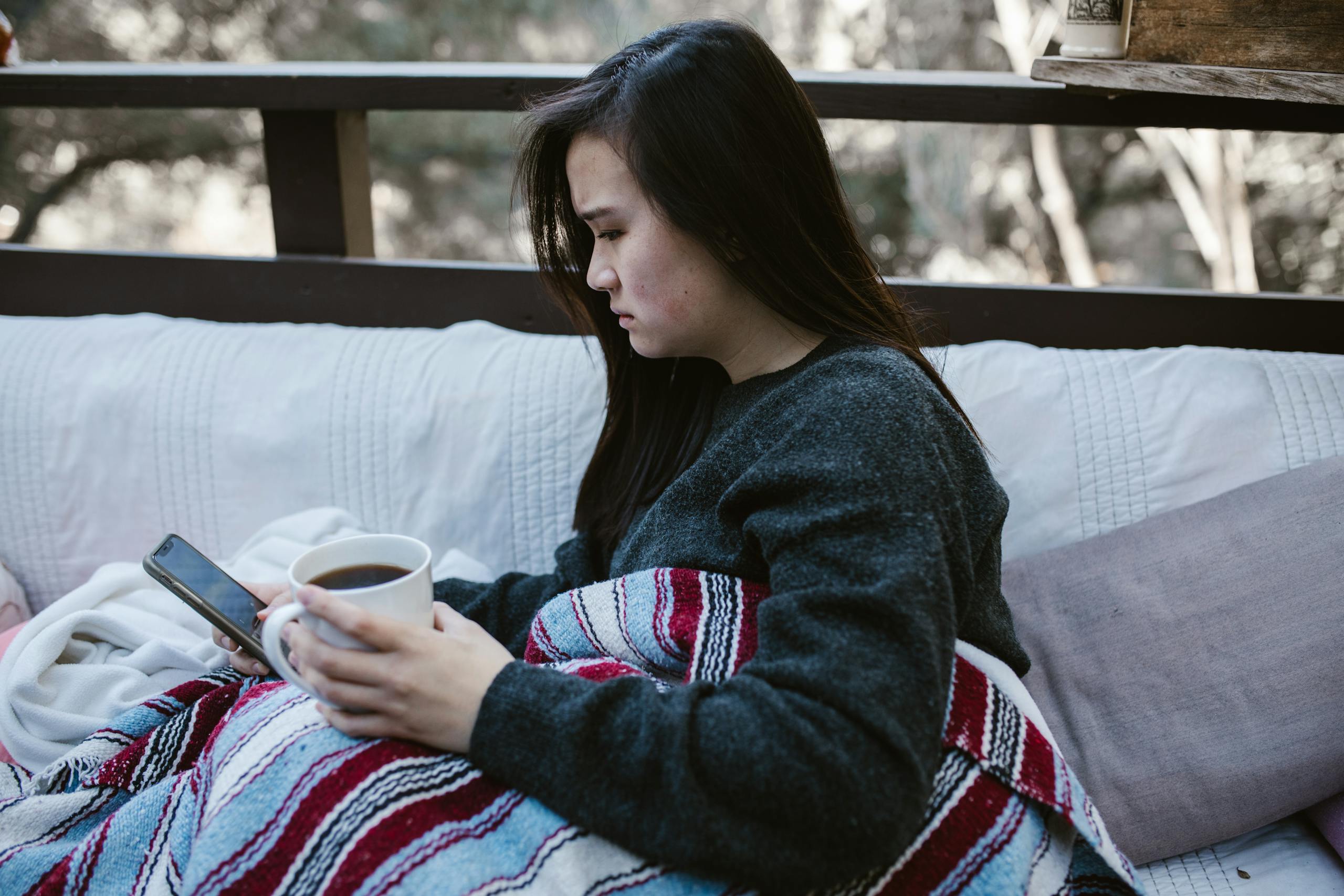 A woman enjoys a quiet moment with coffee and a phone on a cozy outdoor couch.
