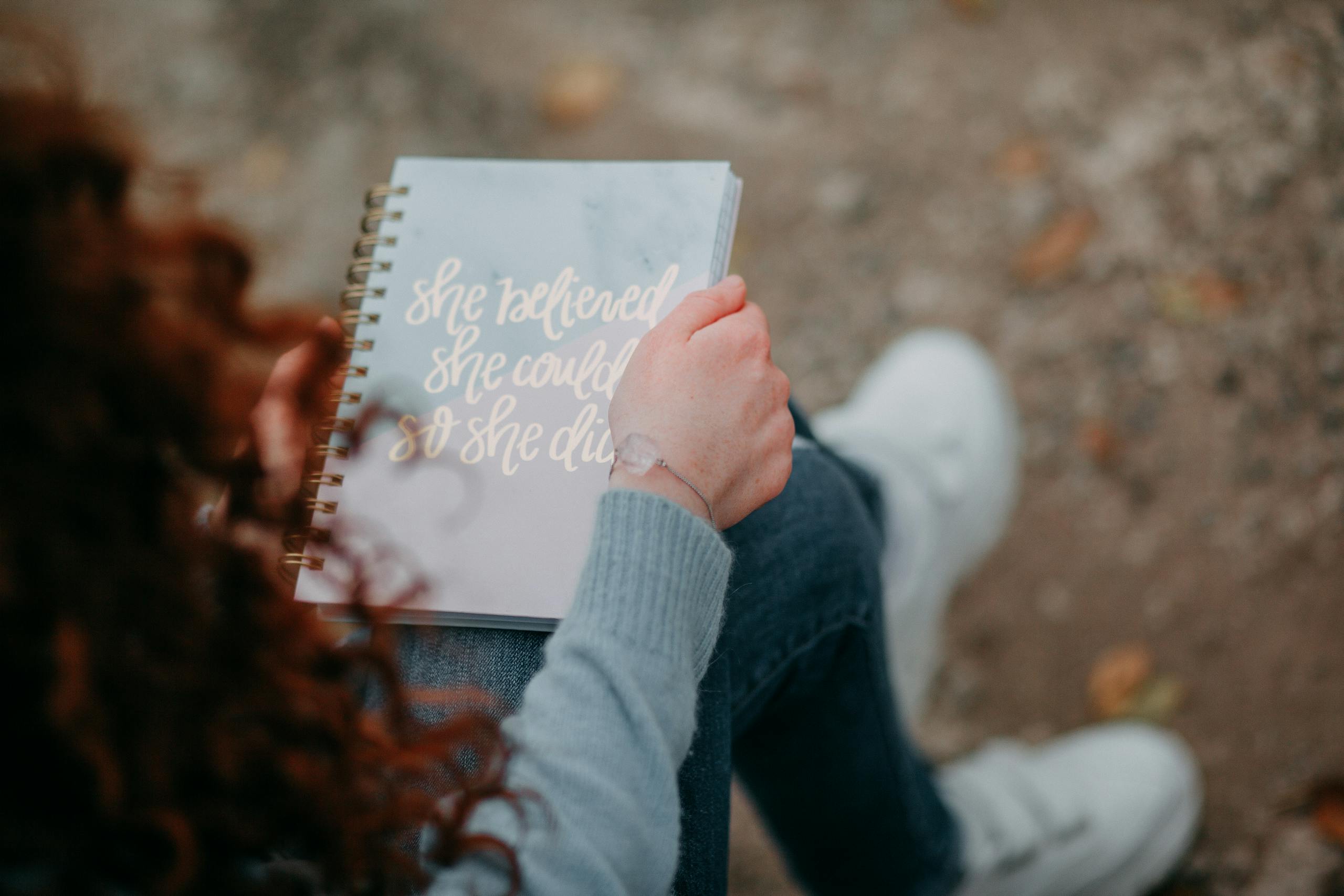 A woman holds a notebook with an inspirational quote, sitting outdoors.