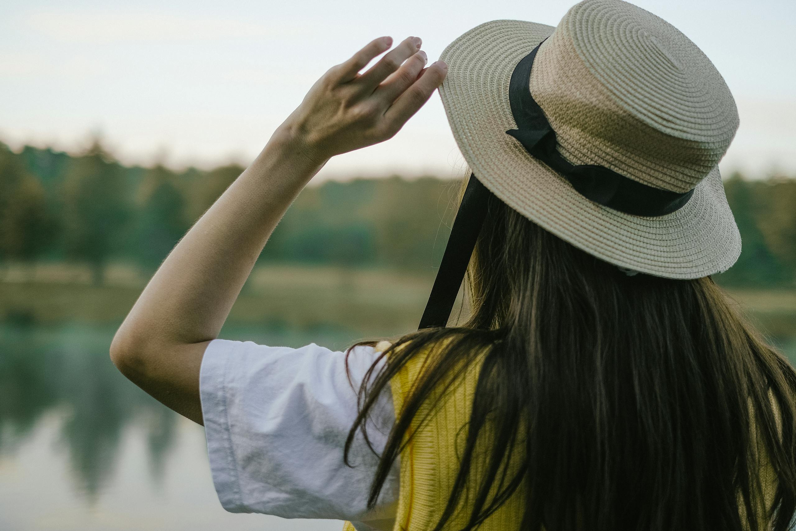 A woman in a straw hat enjoys a serene outdoor view by a lake, capturing a peaceful moment.