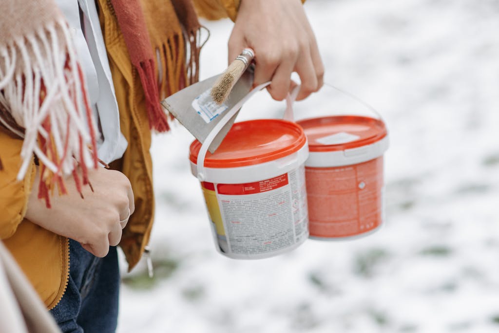 A woman in a yellow jacket holds paint buckets and a brush on a snow-covered day outdoors.