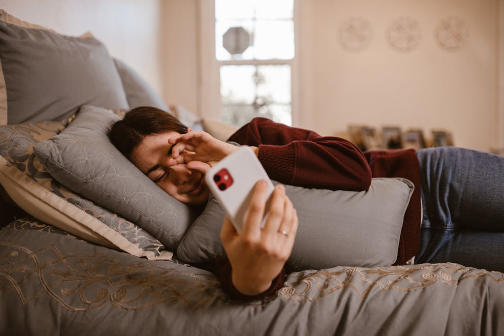 A woman lies in bed, browsing her smartphone, surrounded by cozy pillows in a warm, indoor setting.