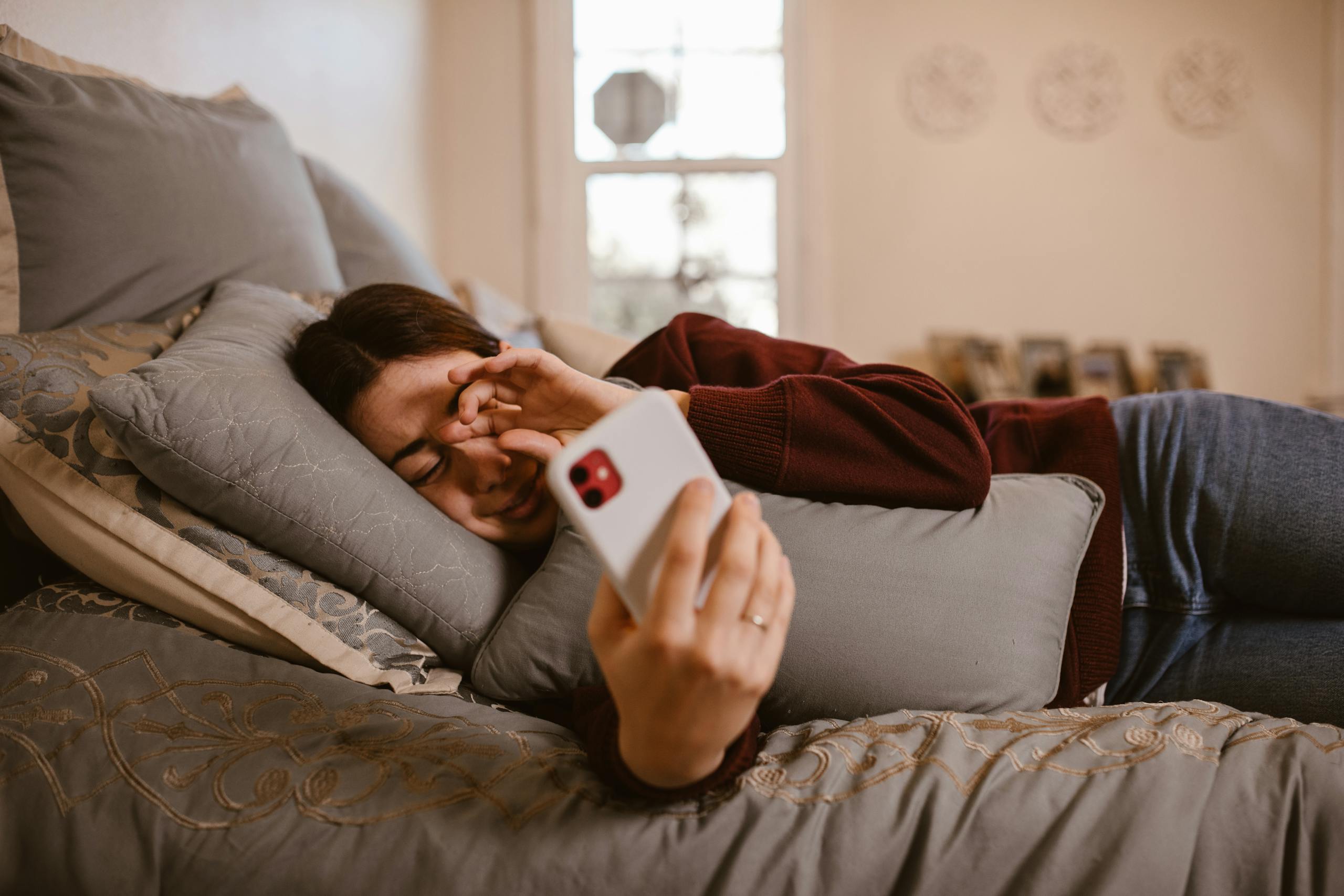 A woman lies in bed, browsing her smartphone, surrounded by cozy pillows in a warm, indoor setting.