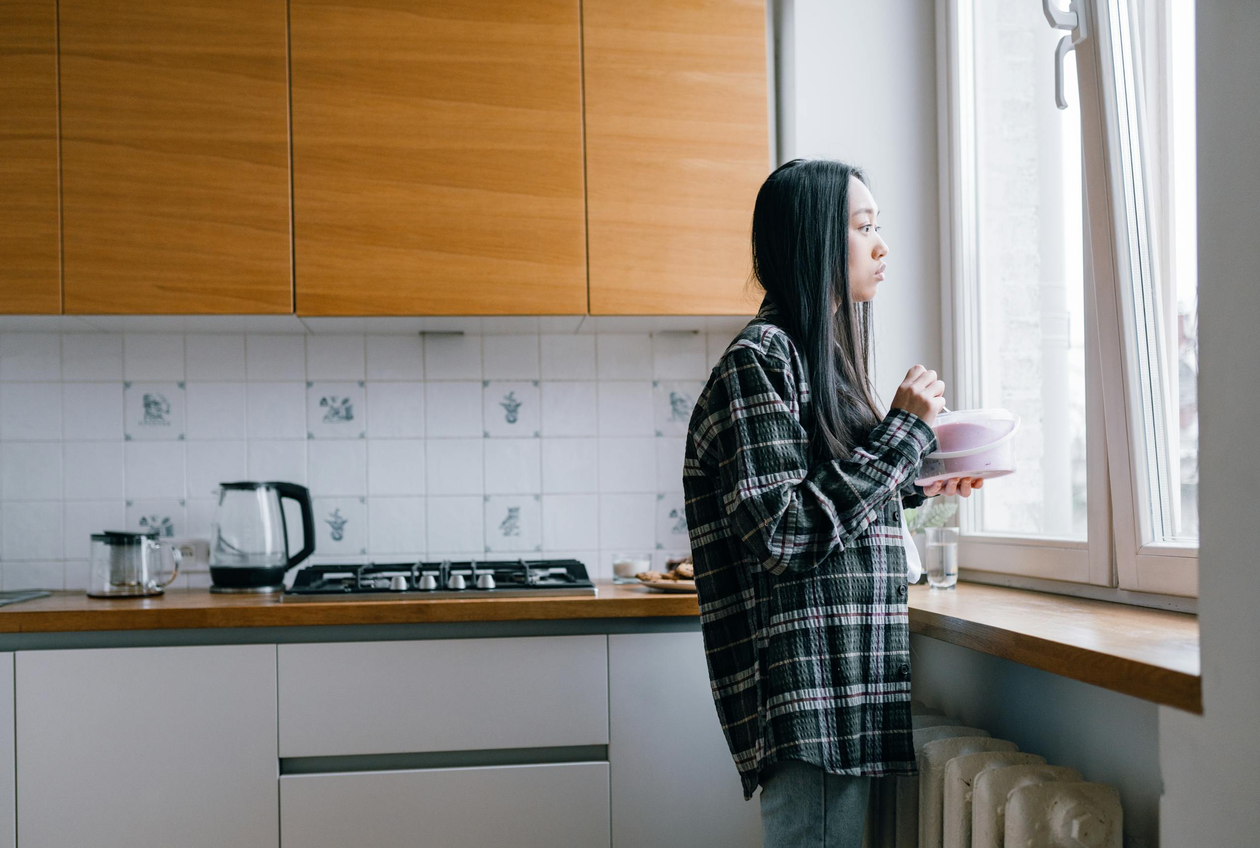 A woman stands in her kitchen, thoughtfully gazing out the window, holding a bowl.