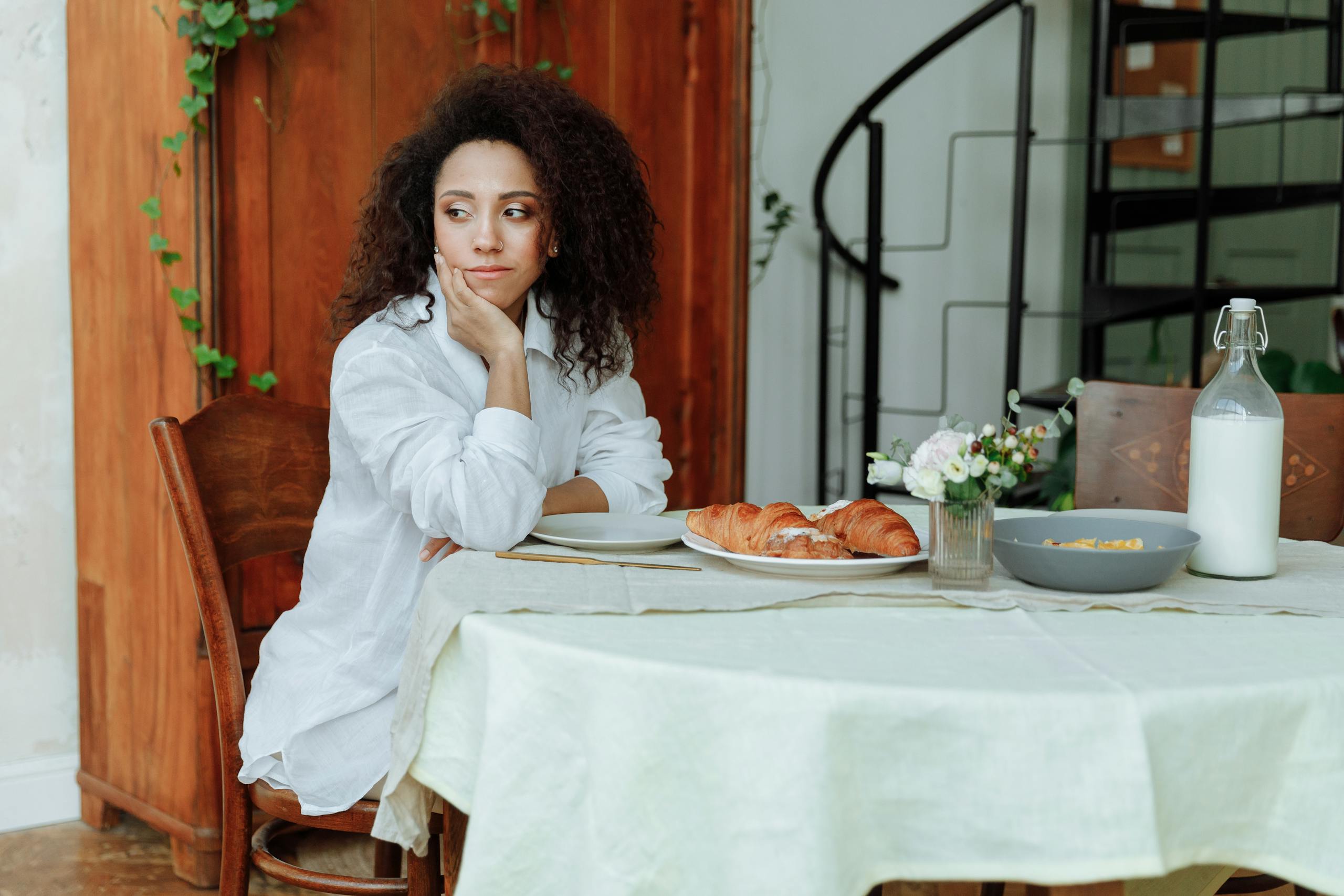 A woman with curly hair sits at a breakfast table with croissants and milk.