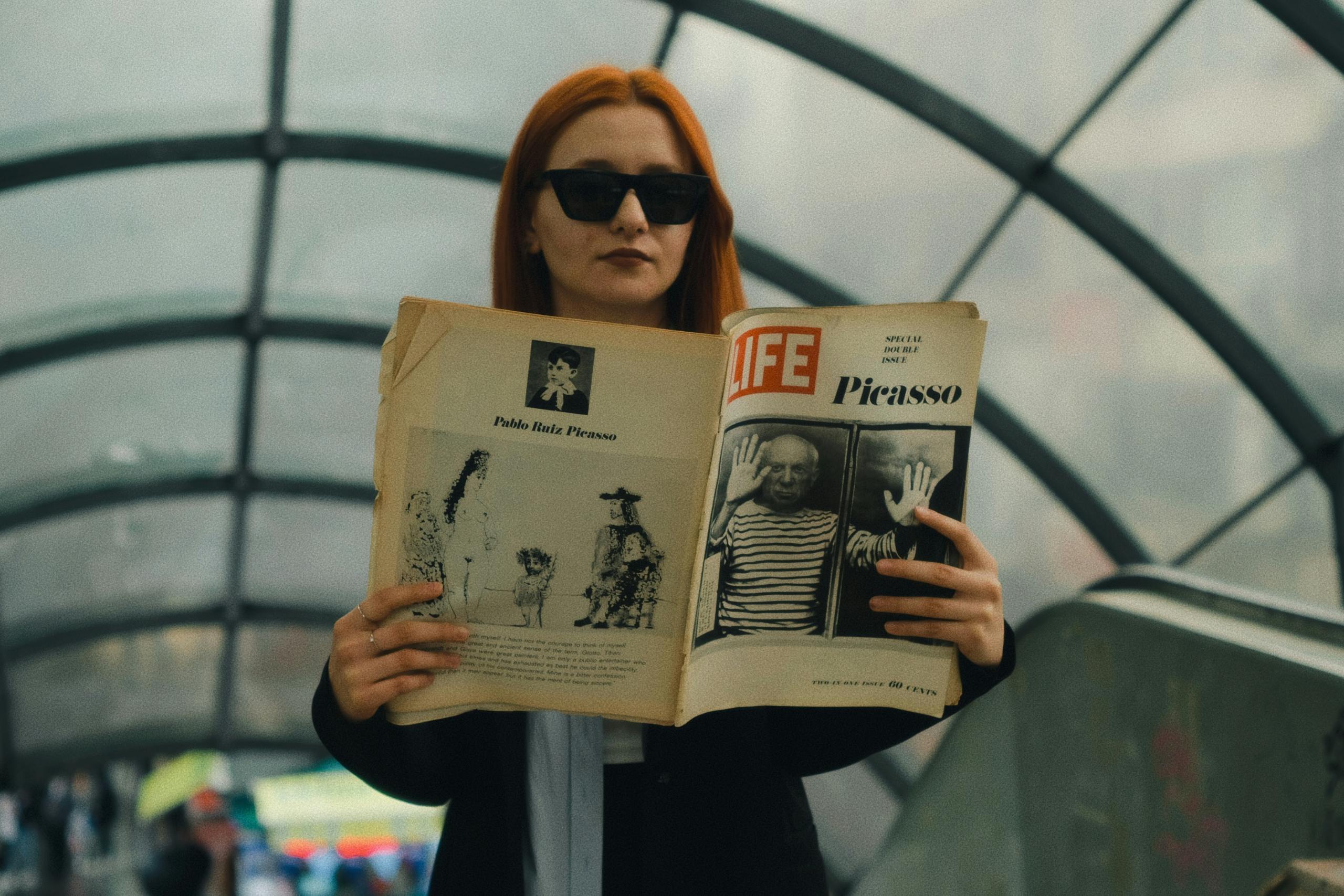 A woman with red hair and sunglasses reads a Life magazine about Picasso in an urban subway setting.