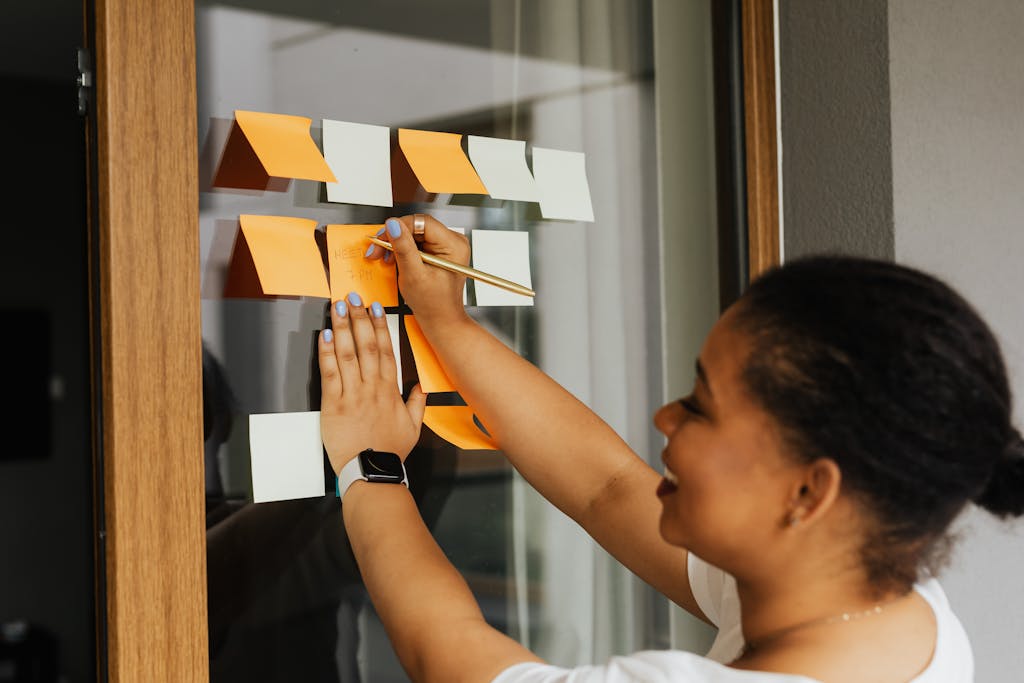 A woman writes notes on a window using colorful sticky notes, emphasizing planning and creativity.