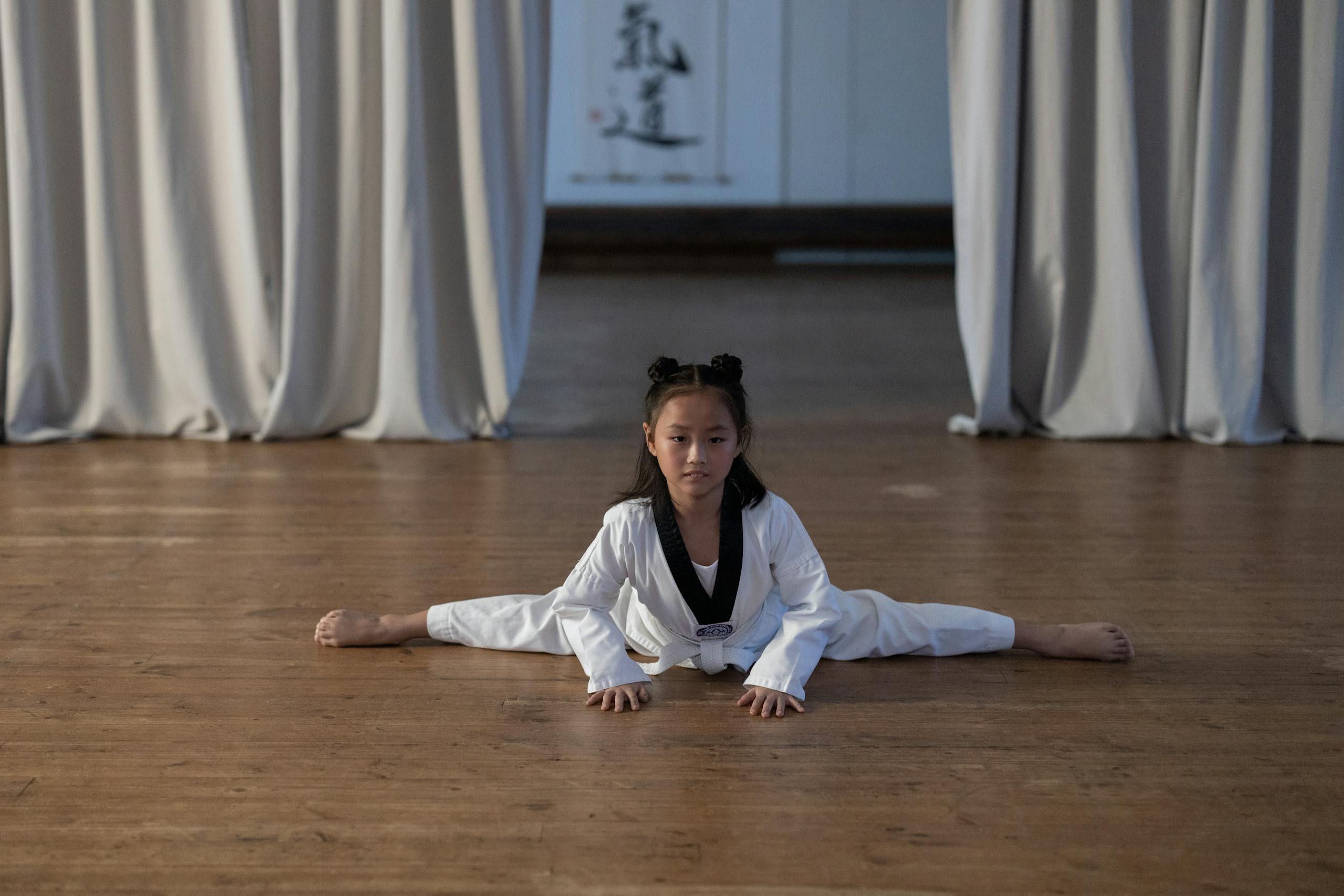 A young girl in a dobok performing a split on a wooden floor during a taekwondo practice indoors.
