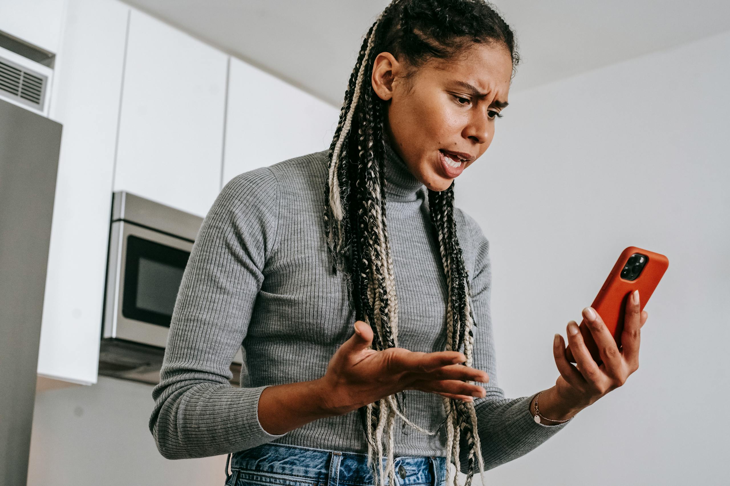 A young woman expressing frustration during a phone call in her kitchen, using a smartphone.