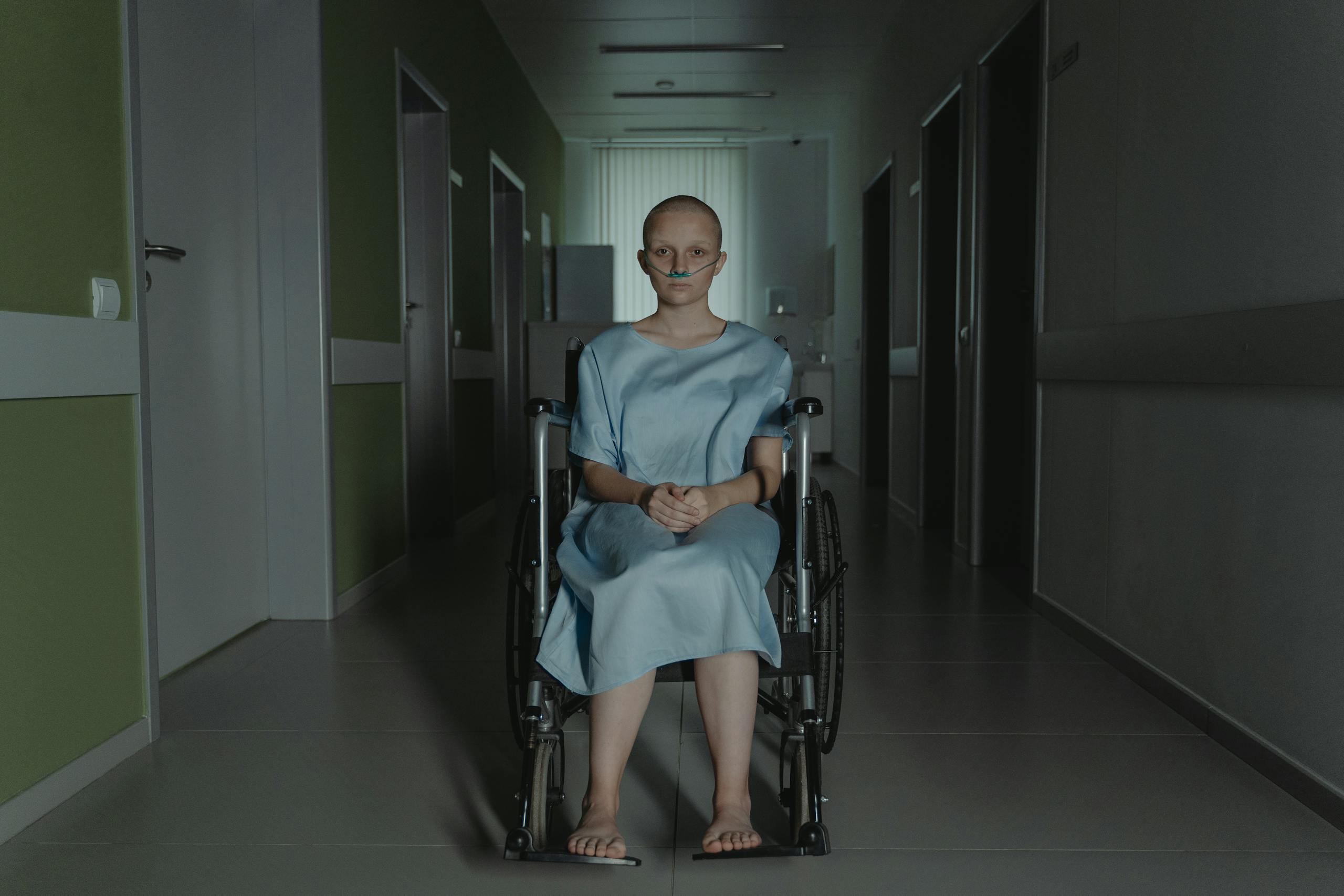 A young woman in a wheelchair sits in a hospital corridor wearing a gown, evoking themes of health and resilience.