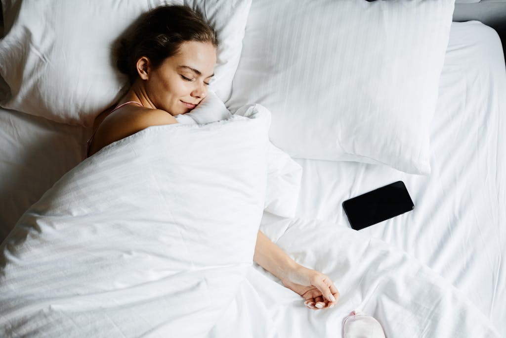 Stop Believing These 10 Sleep Myths, They’re Wrecking Your Rest 3 A young woman sleeps comfortably in bed with a smartphone beside her, wrapped in white sheets.