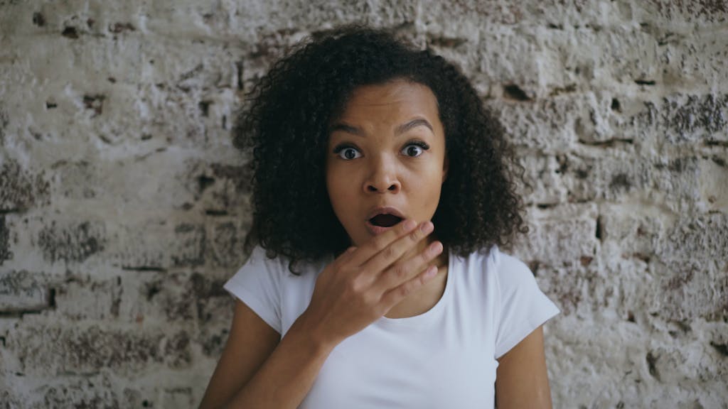 Amazed woman covering her mouth while standing by a rustic brick wall indoors.