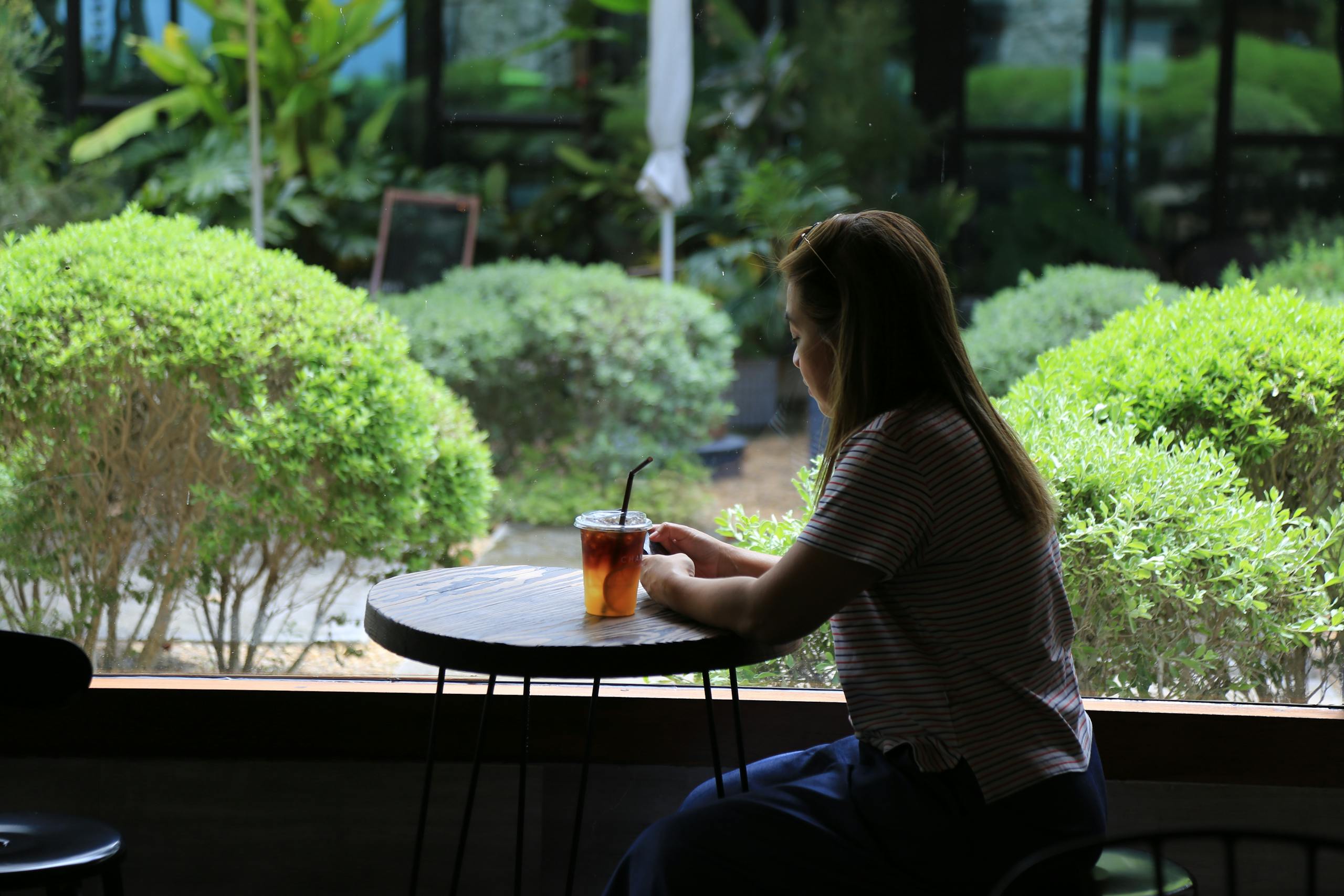 An Asian woman sits in a garden café, enjoying a cold drink on a summer day.