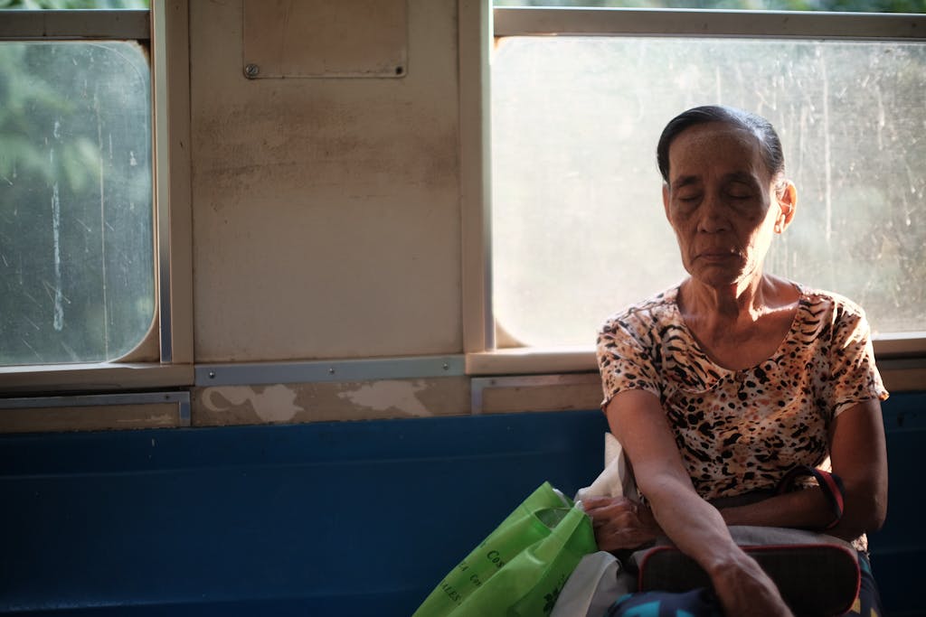 An elderly woman with closed eyes rests peacefully while sitting inside a sunlit bus.