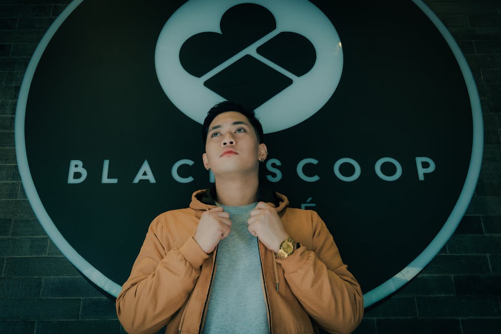 Asian man wearing a brown jacket stands confidently near Black Scoop Cafe signage, low angle view.