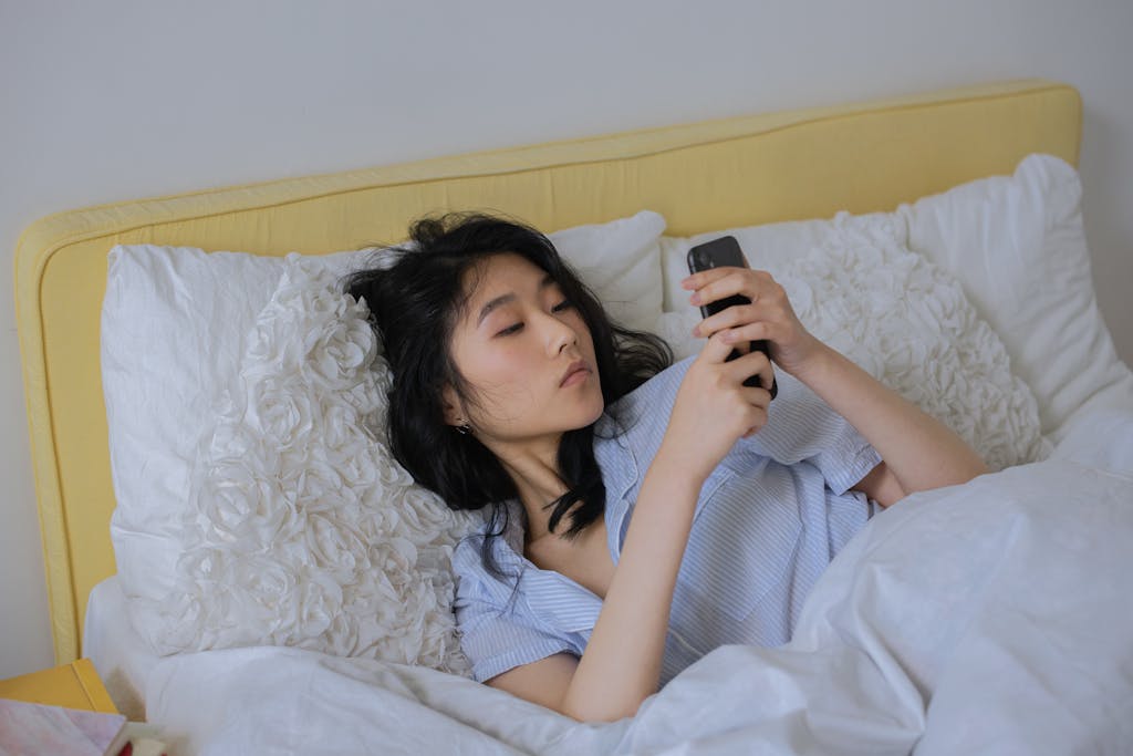 Asian woman comfortably lying in bed, using her phone in a serene bedroom setting.