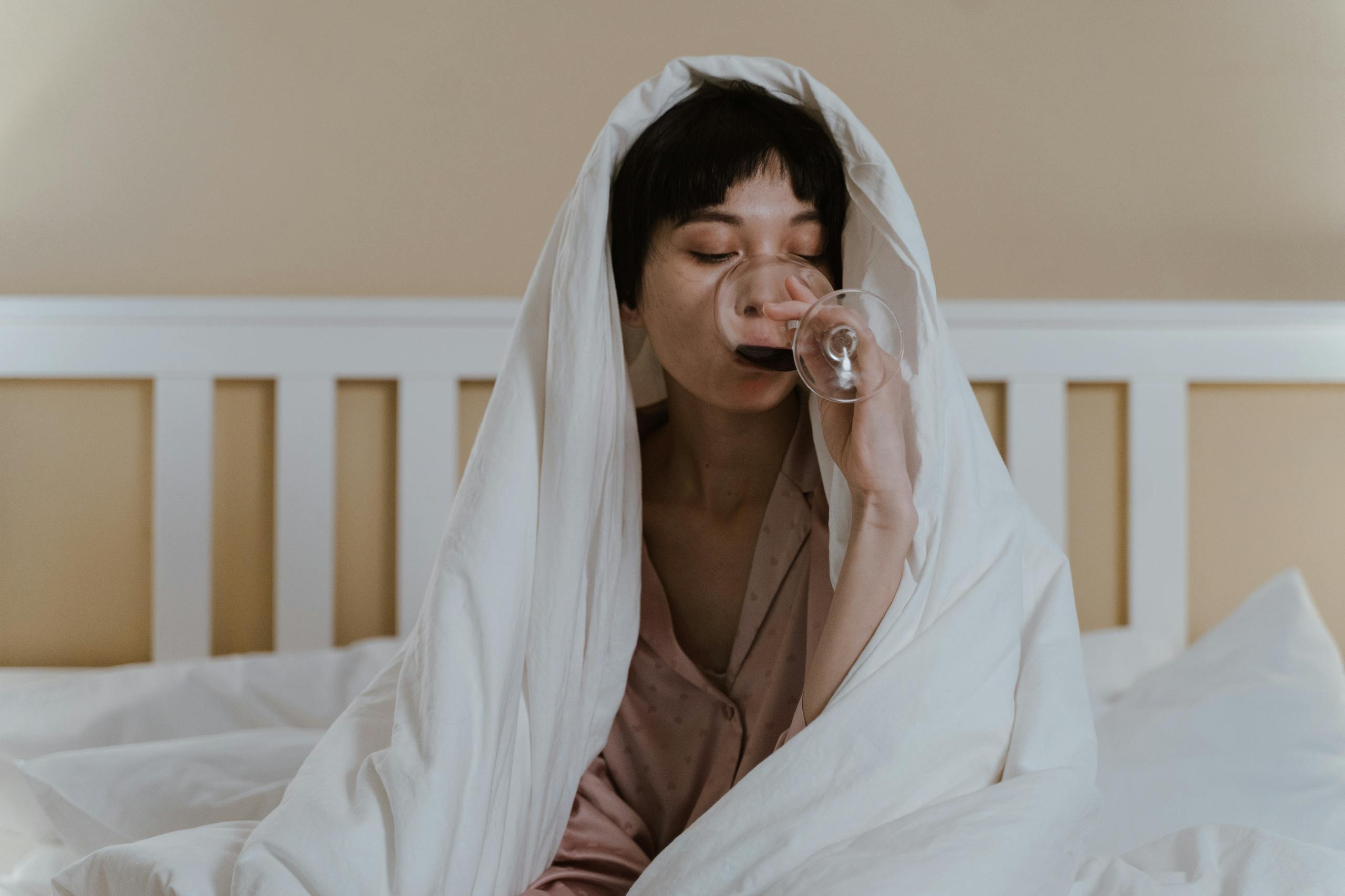 Asian woman relaxing in bed, wrapped in a blanket, enjoying a glass of wine.
