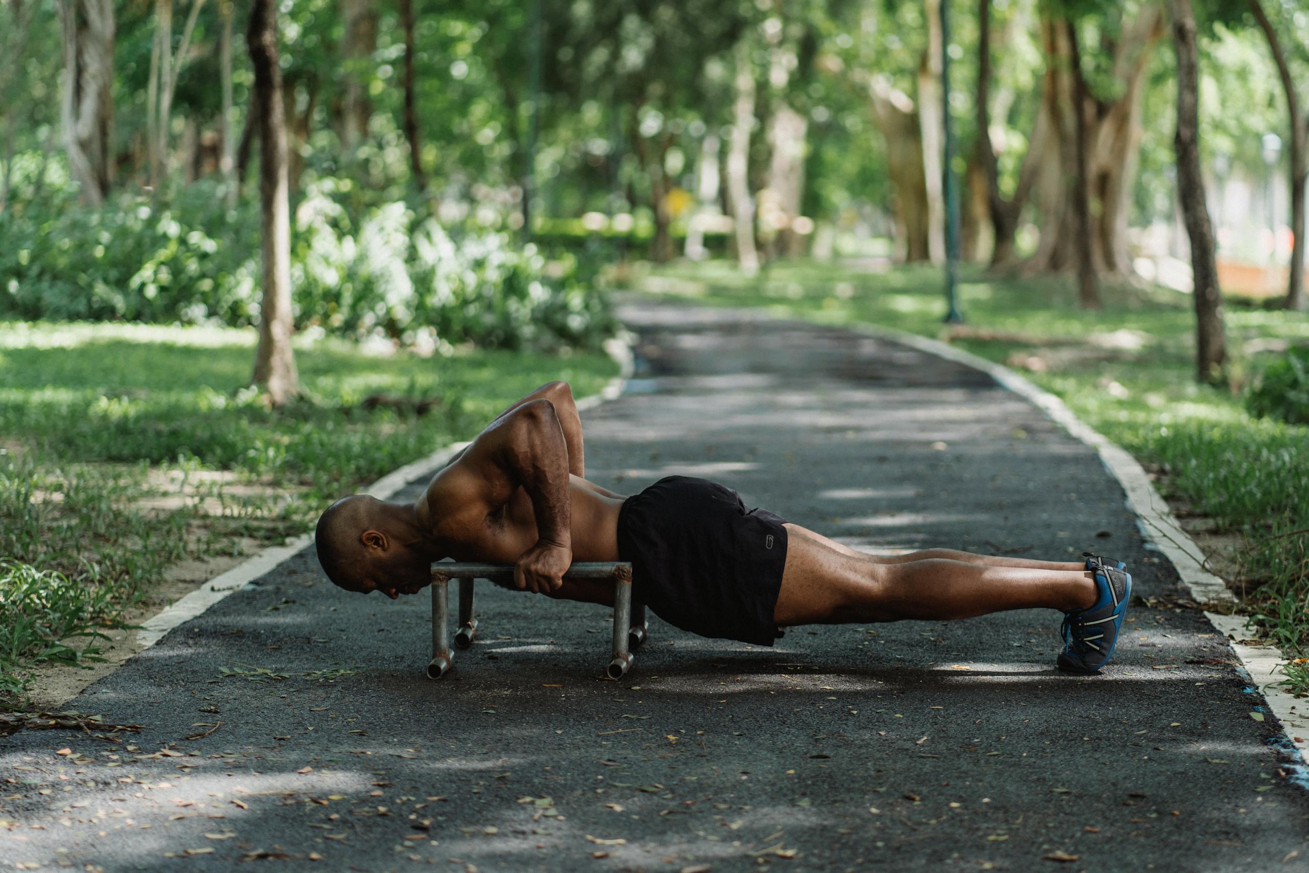 Athletic man performing push ups outdoors on a park pathway surrounded by green trees.