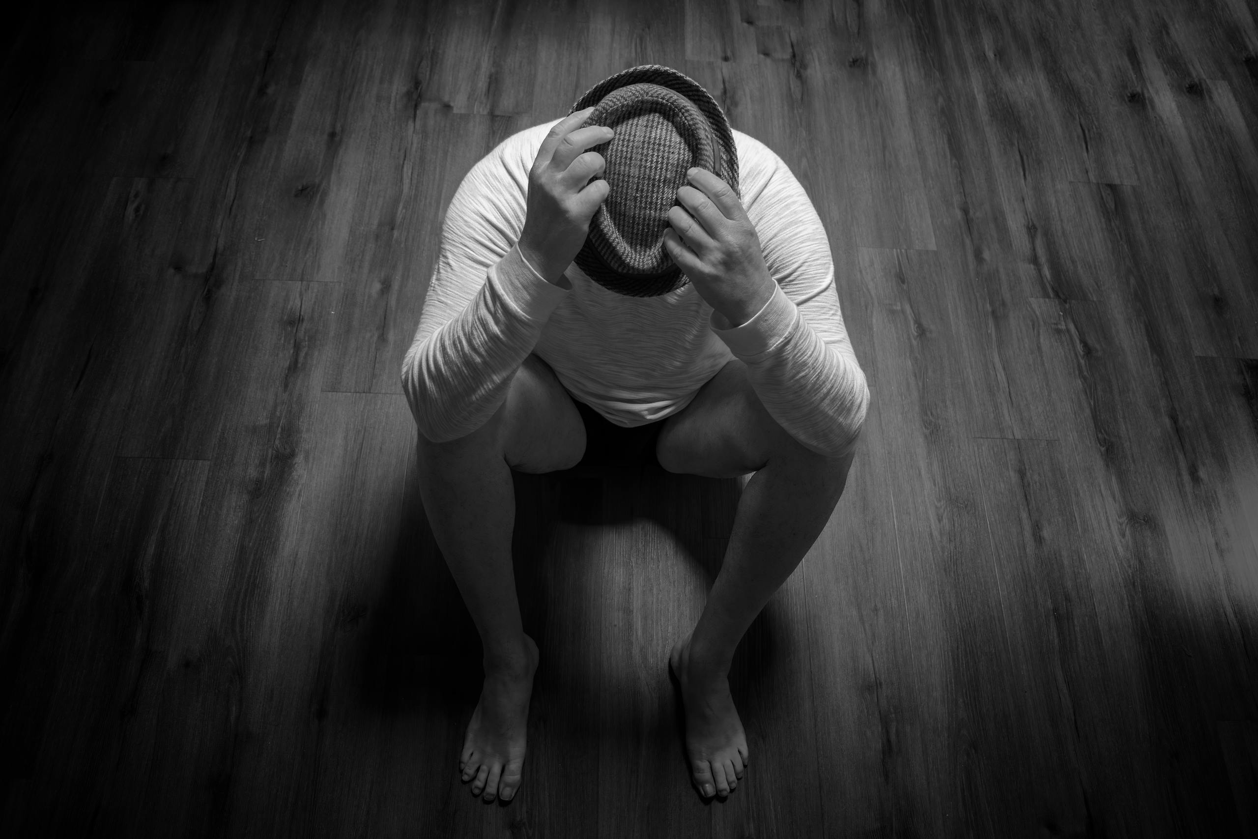 Black and white photo of a person sitting with a hat on a wooden floor, conveying sadness.