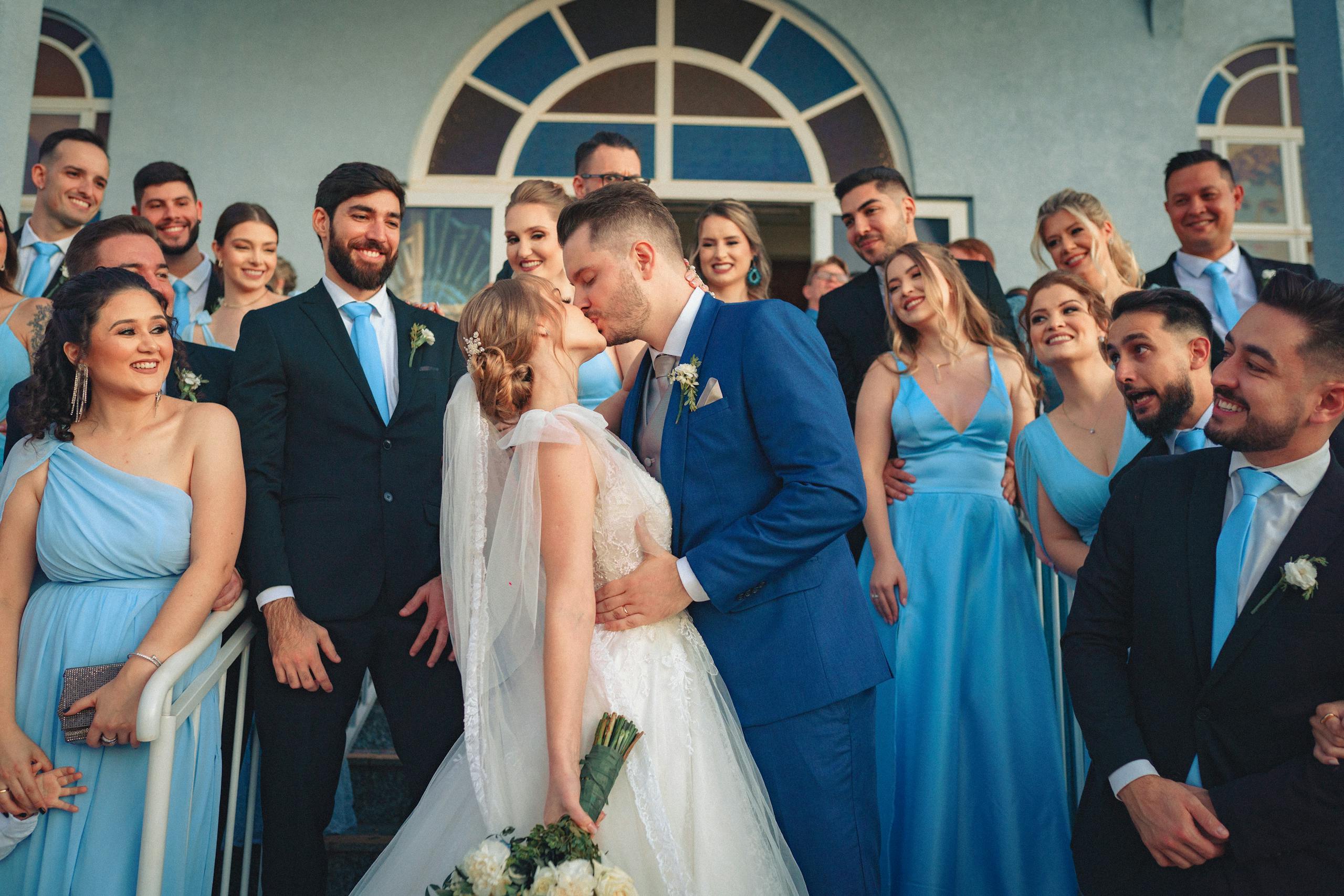 Bride and groom kiss surrounded by joyful wedding party in blue attire.