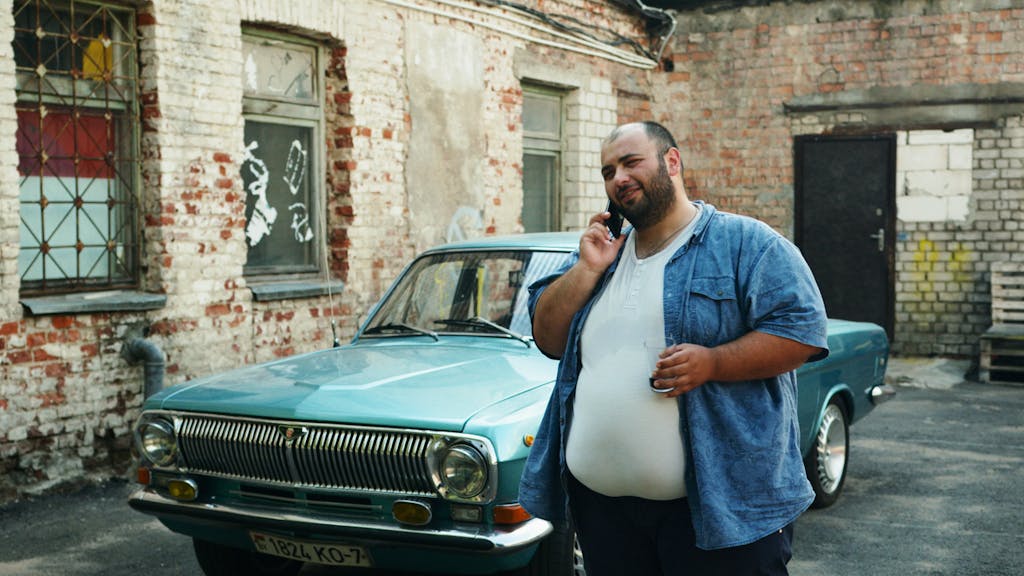 Casually dressed man talking on the phone next to a vintage car in an urban setting.