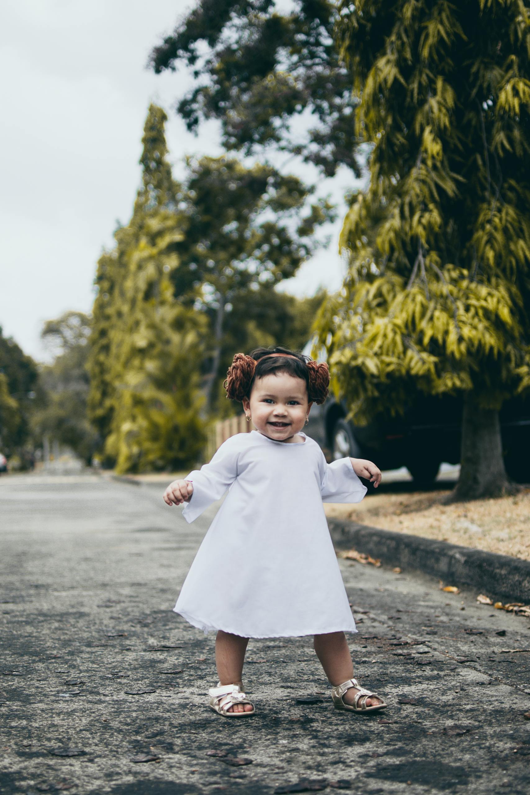 Charming toddler in a white dress smiling and having fun outdoors on a sunny day.