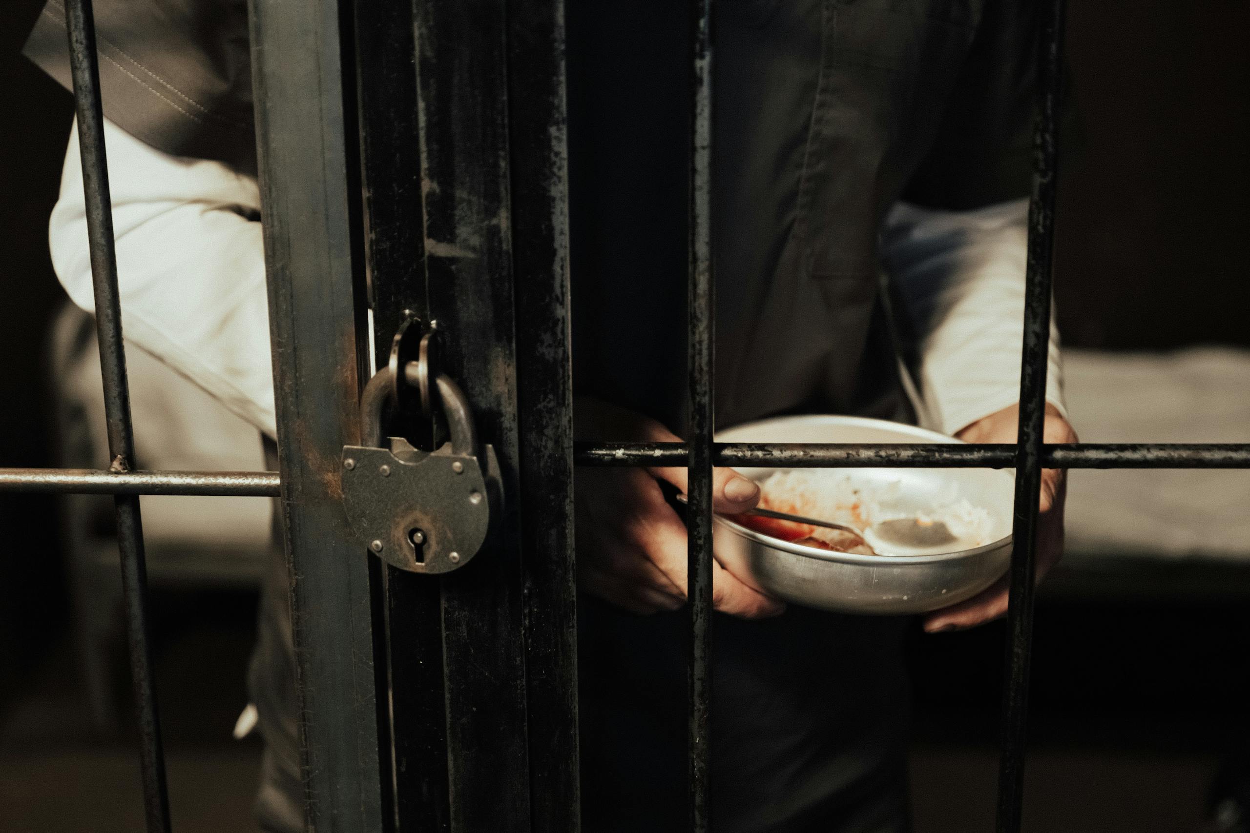 Close-up of a prisoner holding a bowl behind iron bars, illustrating confinement.