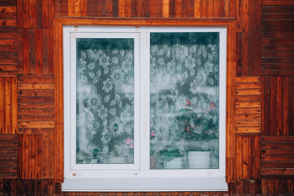 Avoid These 8 Heat Wave Mistakes Before They Harm Your Health 1 Close-up of a wooden house exterior with a floral curtain window revealing potted plants.