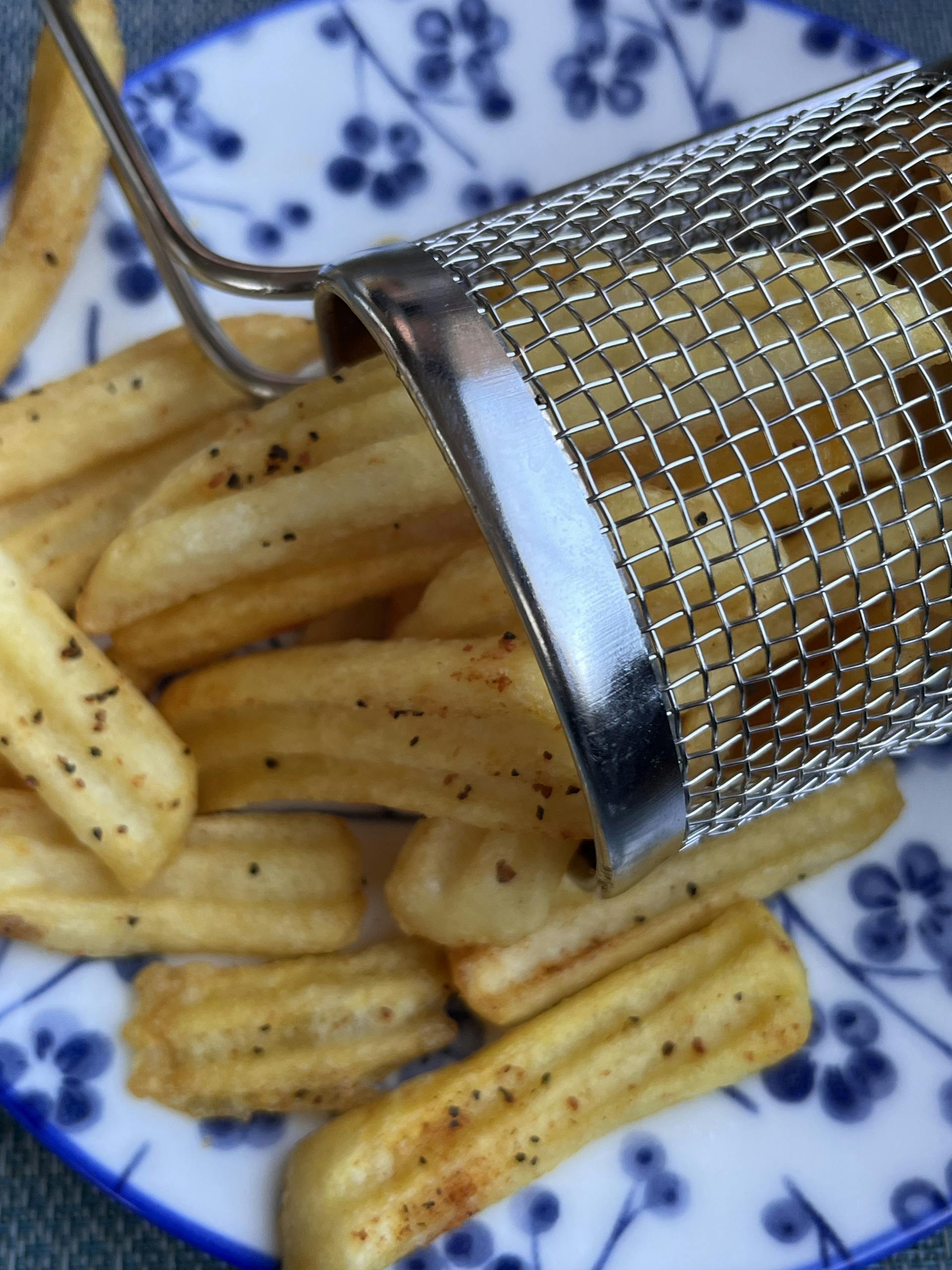 Close-up of crispy seasoned fries in metal basket on floral plate.