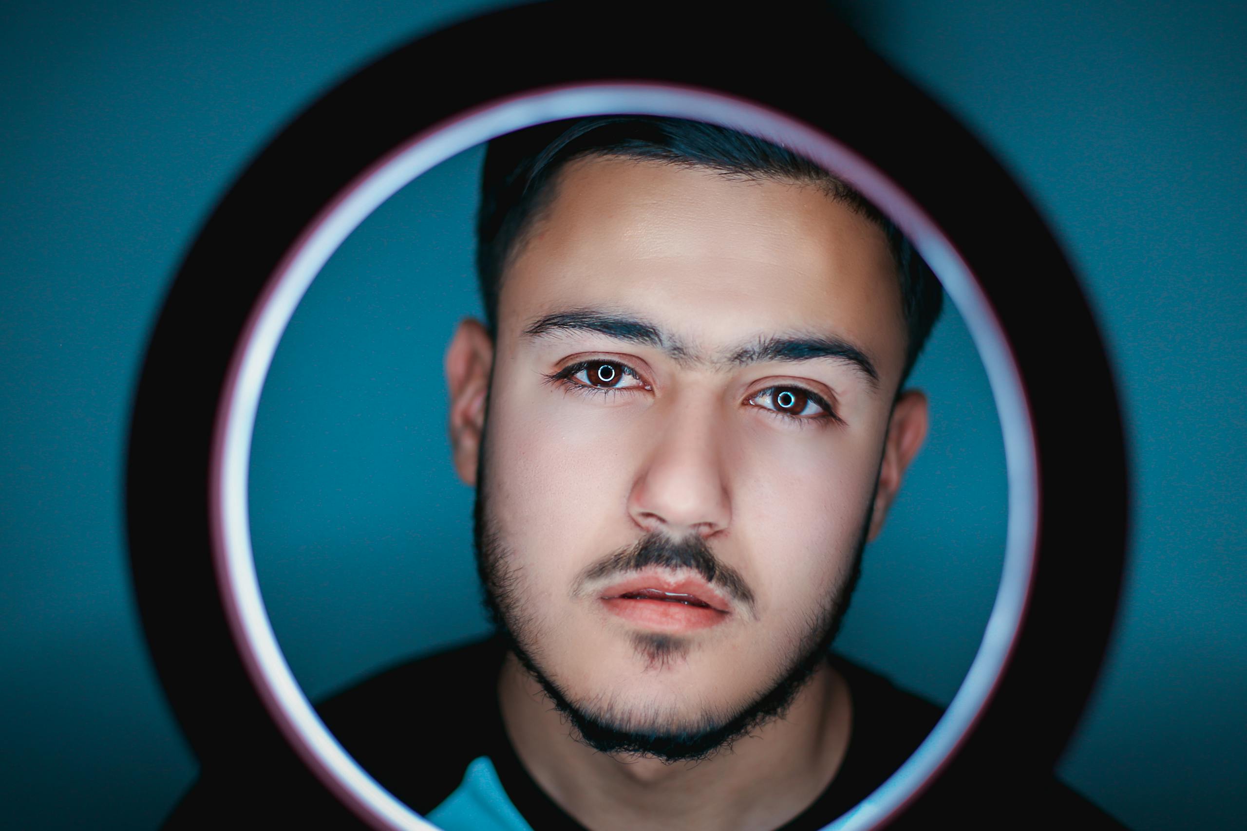 Close-up portrait of a young man with facial hair, framed by a ring light for a modern photography look.
