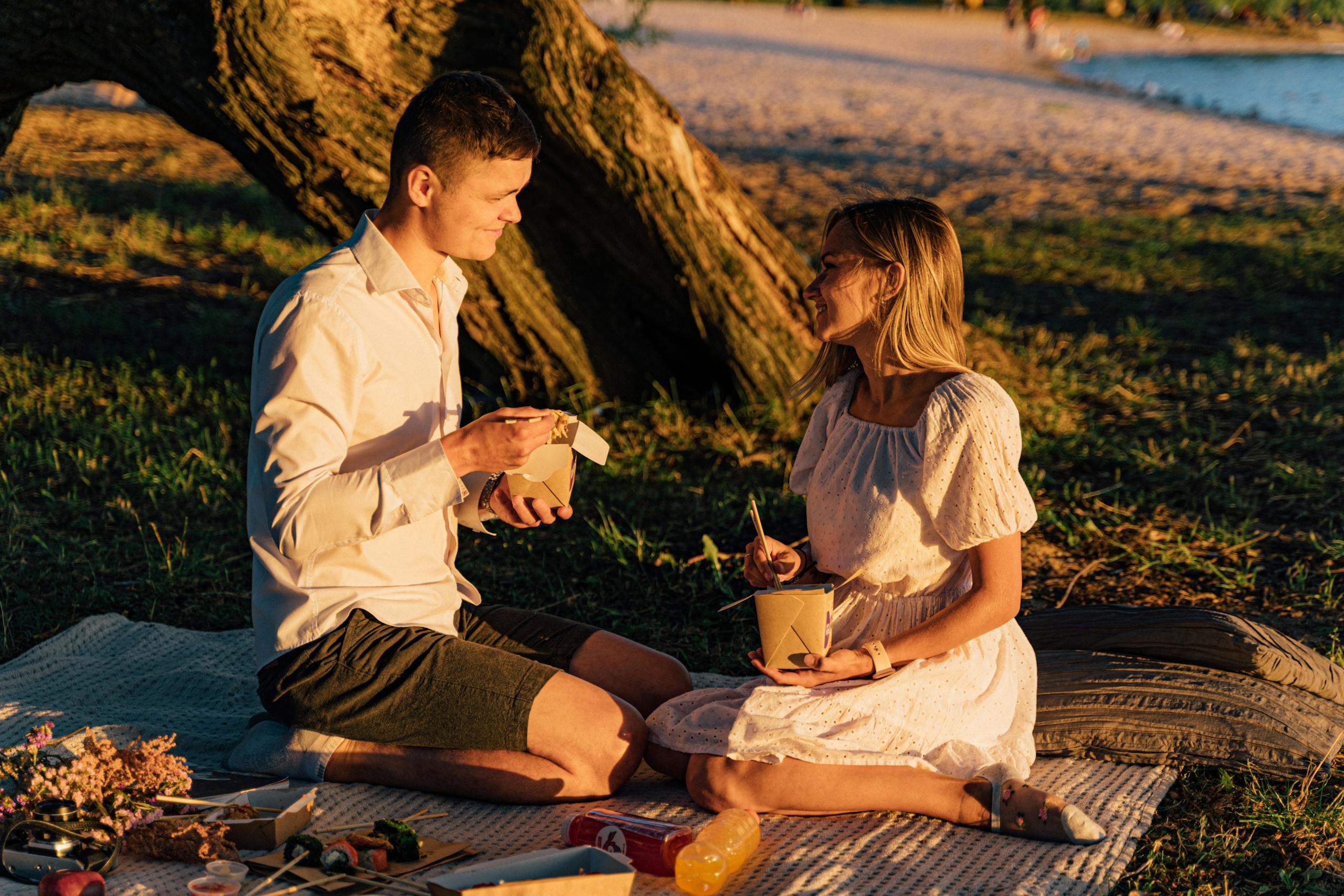 Couple enjoys a sunset picnic with takeout noodles by the beach. Perfect summer evening.