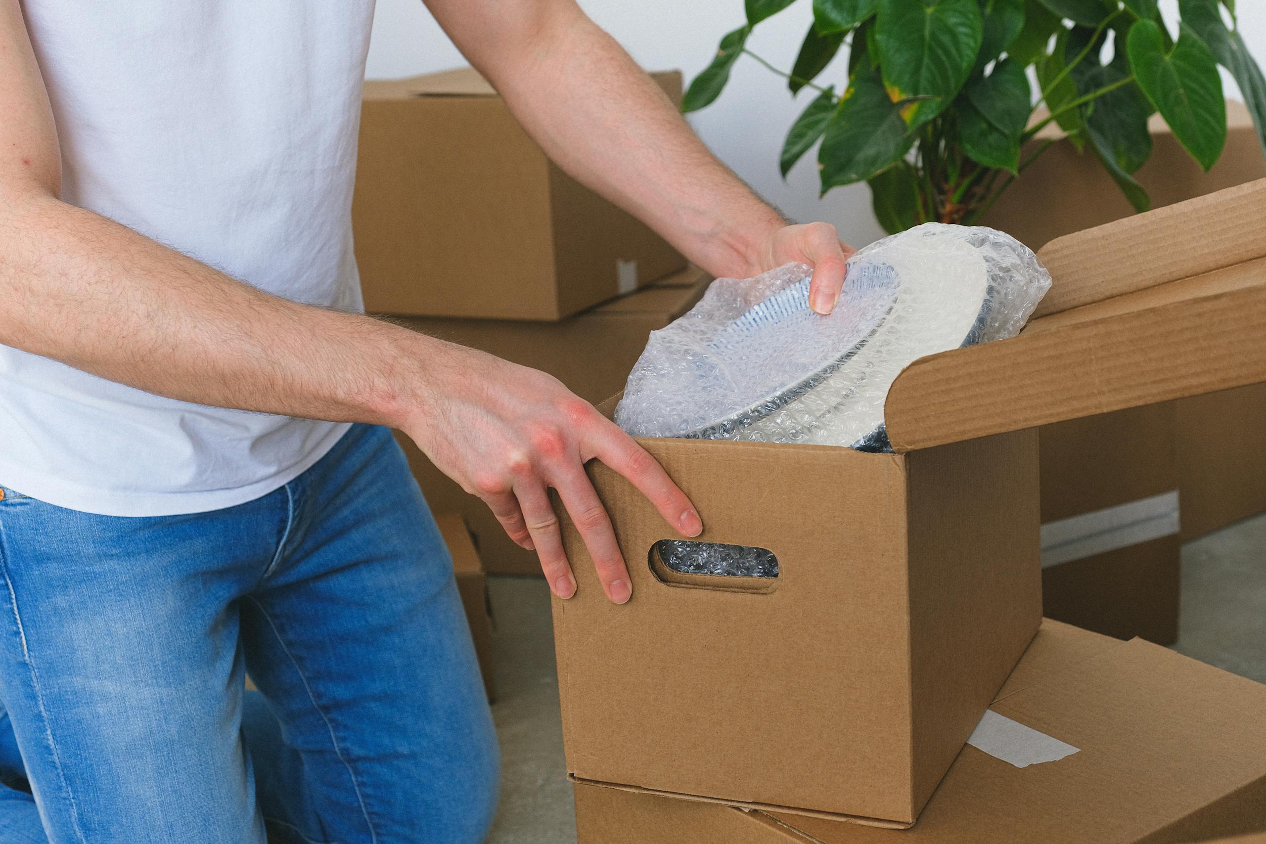 Crop anonymous male in casual clothes packing fragile plates in carton box before moving into new flat in daylight