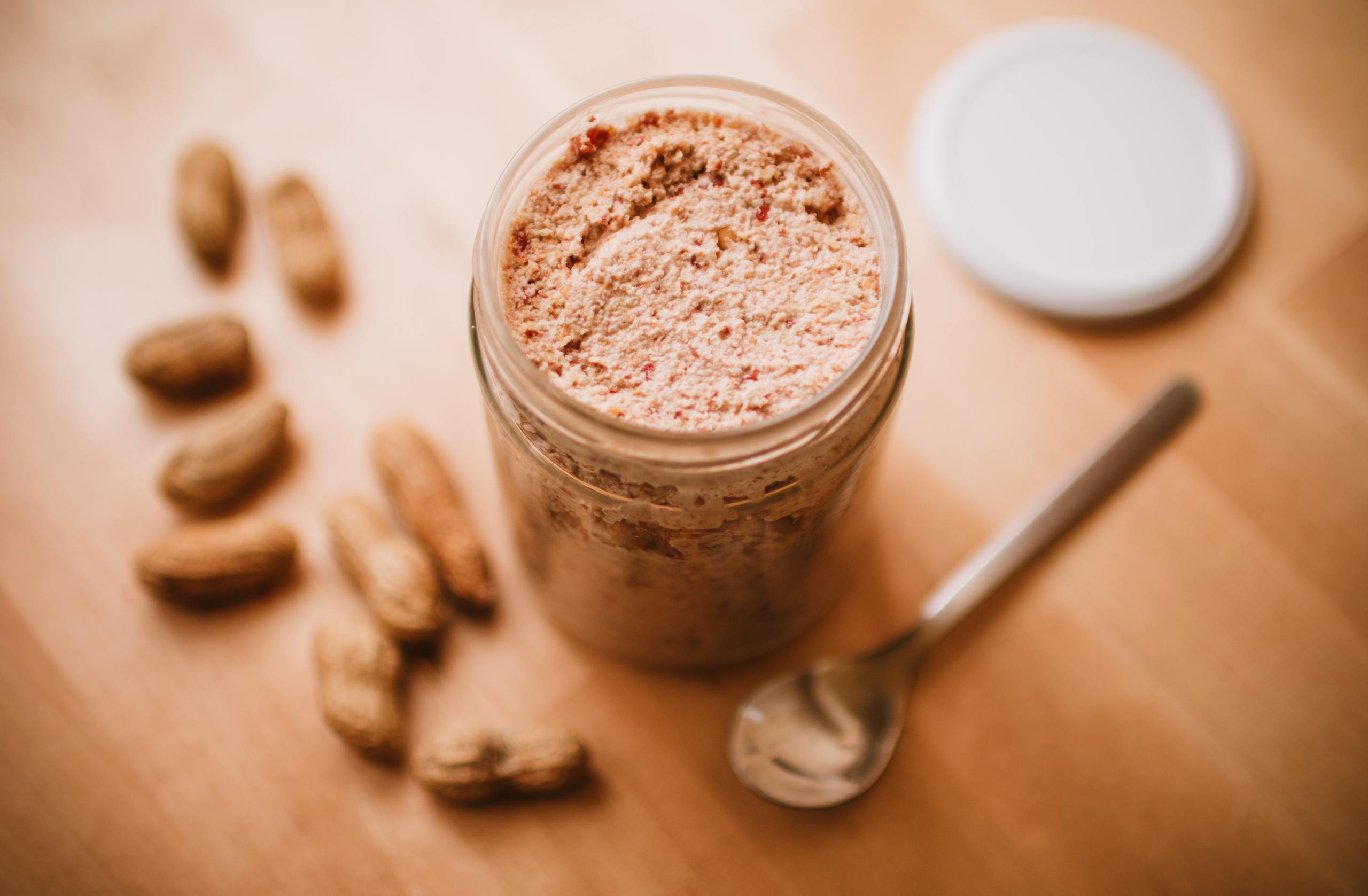 Delicious homemade peanut butter in a glass jar with peanuts and a spoon nearby.